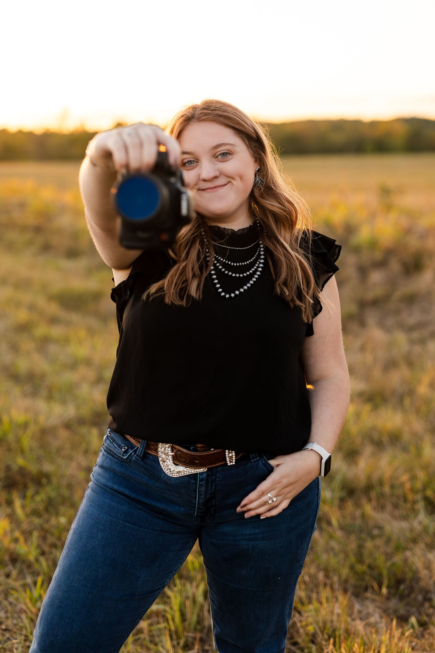 Laura, Female photographer extending camera toward viewer with a smile, standing in a sunlit field at golden hour.