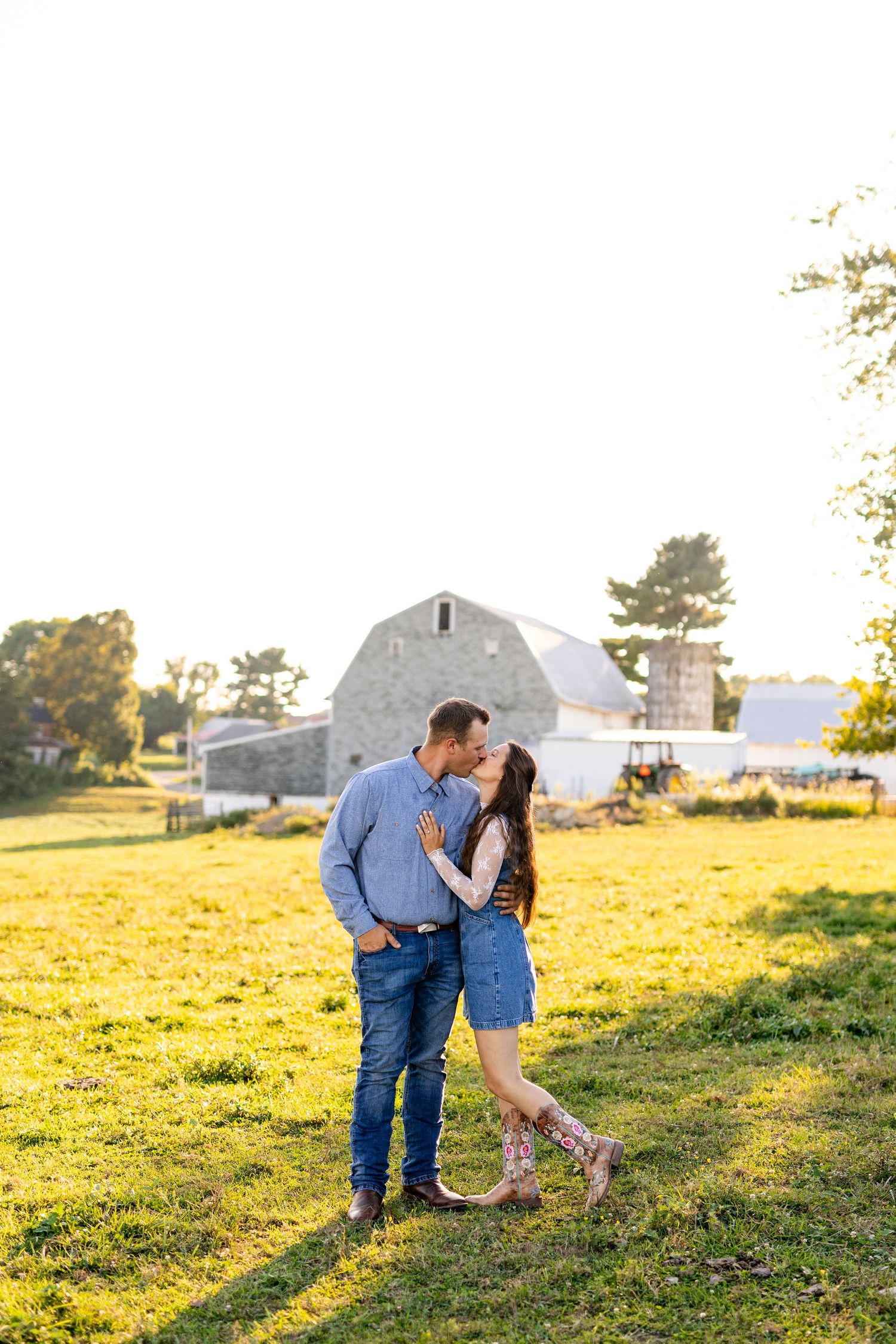 Couple sharing a kiss in a sunlit farm field with a rustic barn backdrop during golden hour engagement session.