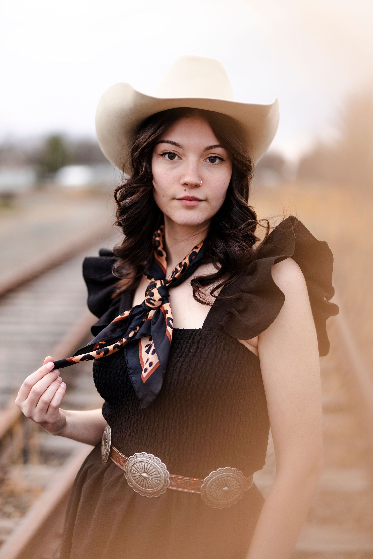 Young woman in white cowboy hat, black ruffled dress, concho belt, and patterned scarf poses on railroad tracks.