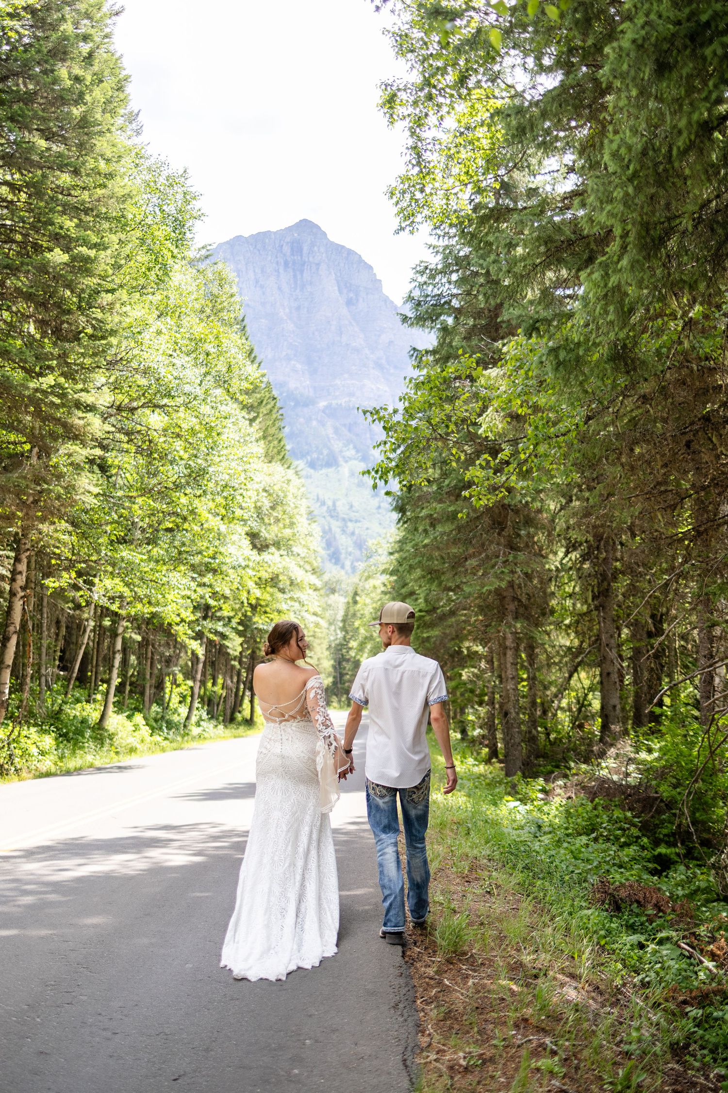 Bride and groom walk hand in hand down a tree-lined road with a mountain vista in the background.