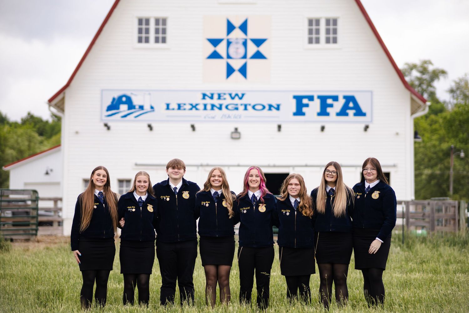 Eight New Lexington FFA students standing together in black jackets before a white barn.