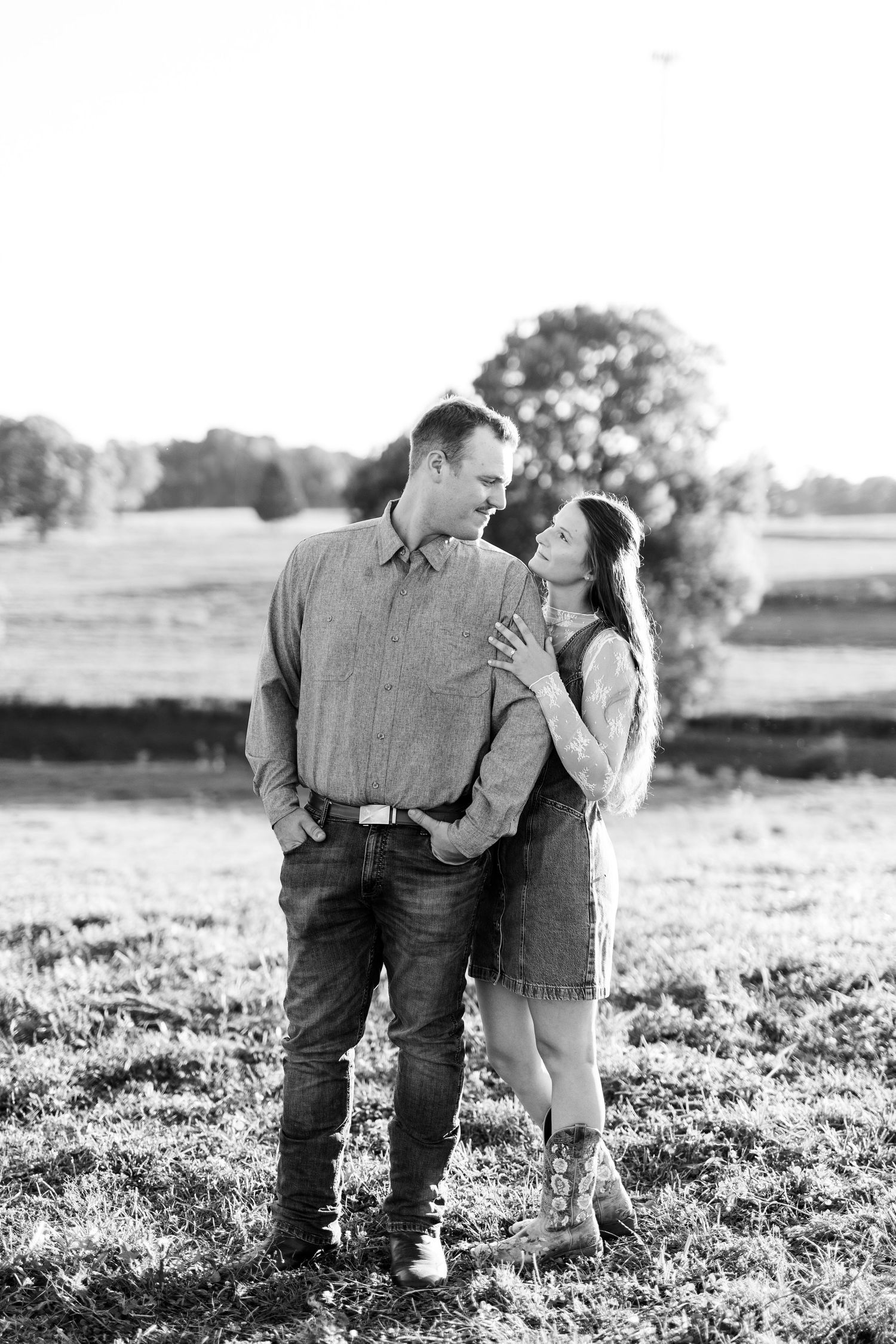 Black and white photo of a couple gazing at each other in a sunny field with trees in the background.