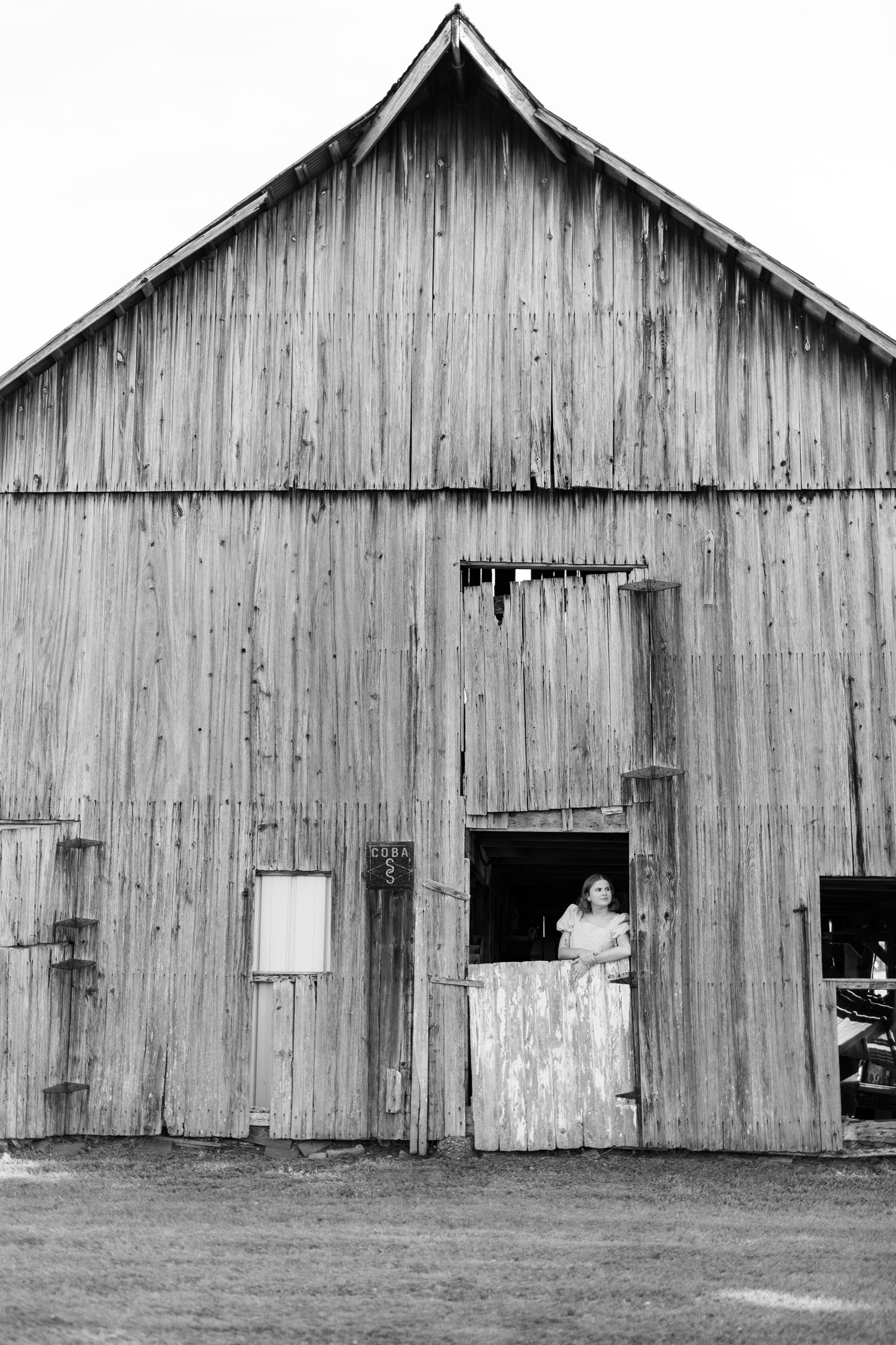 Black and white photo of a weathered wooden barn with a person standing in the open doorway on a rural farm.