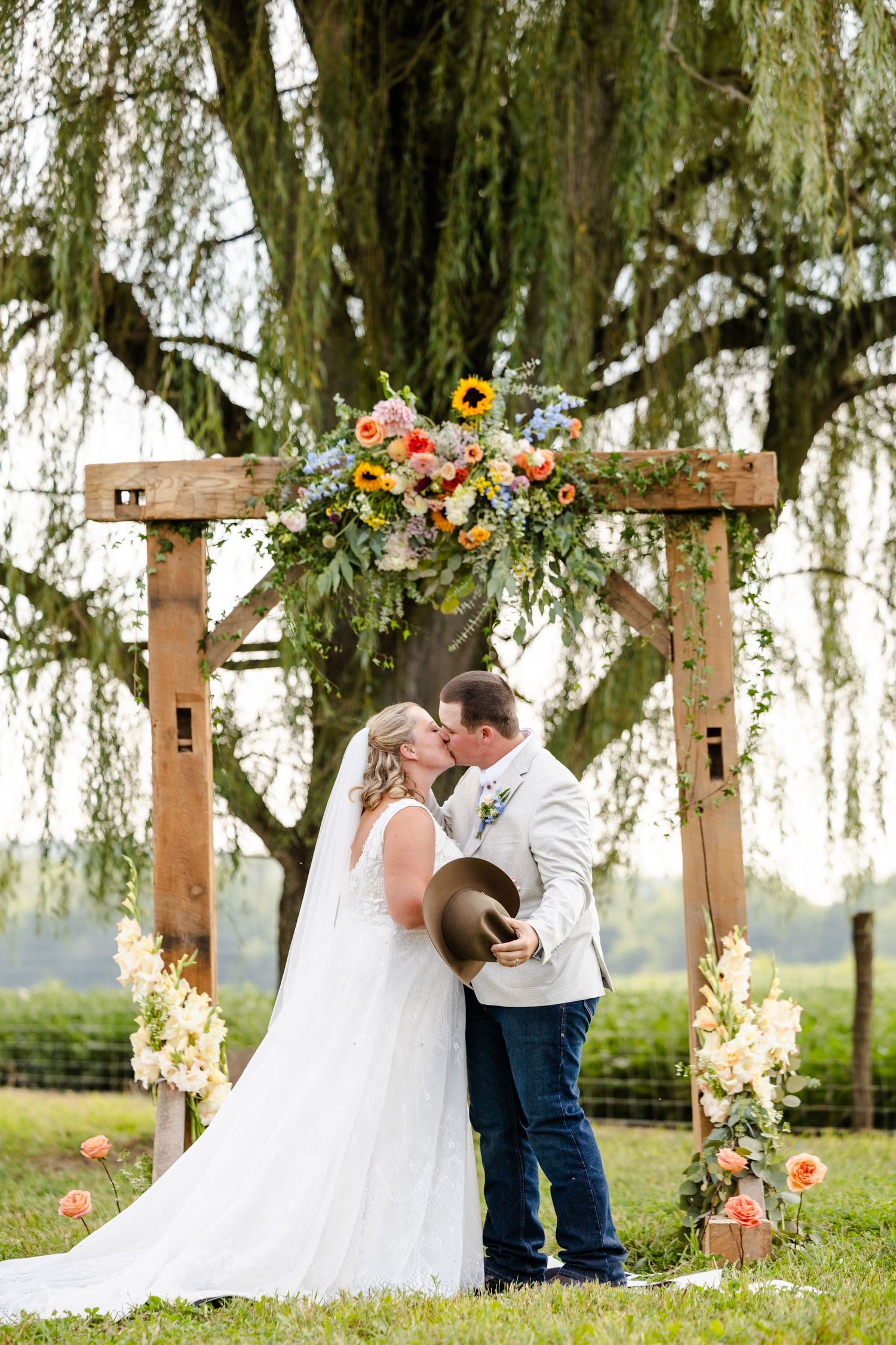 Bride and groom share their first kiss under a sunflower-adorned wooden arch beneath a sprawling weeping willow tree.