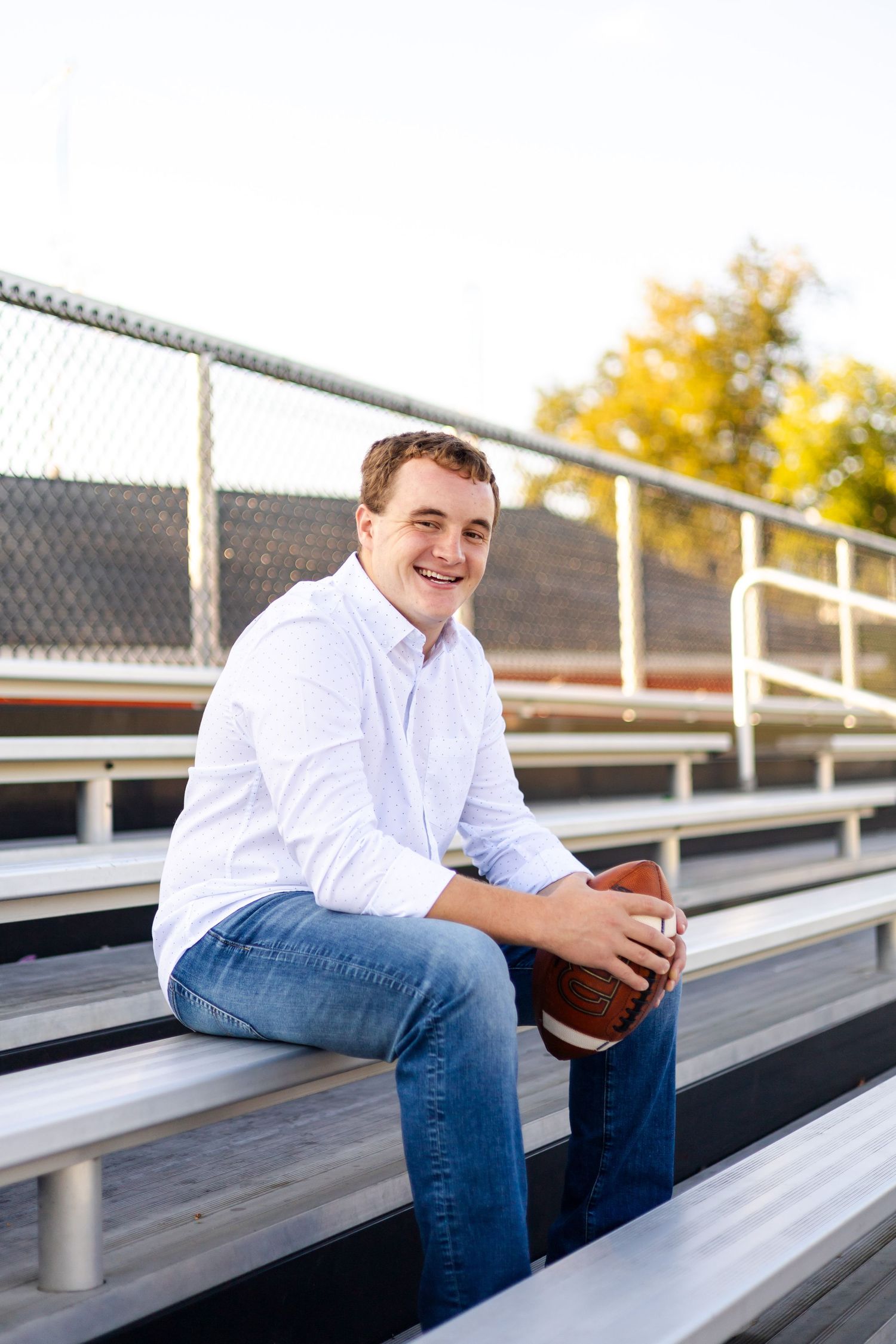 Young man in white shirt and jeans sitting on bleachers holding a football, smiling outdoors.