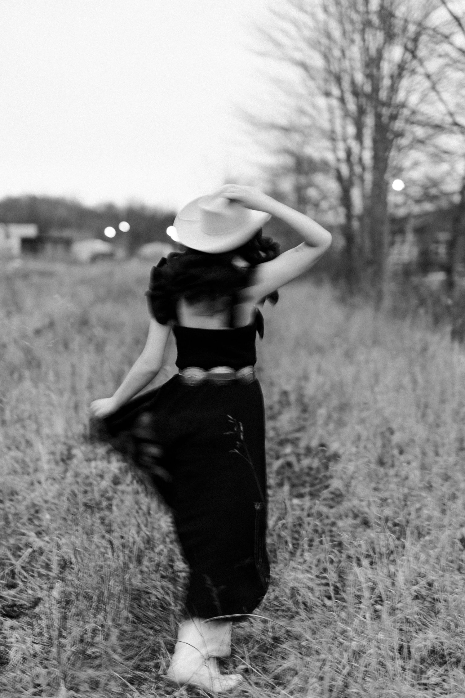Black and white photo of a woman in cowboy hat, crop top, and boots walking through a field.