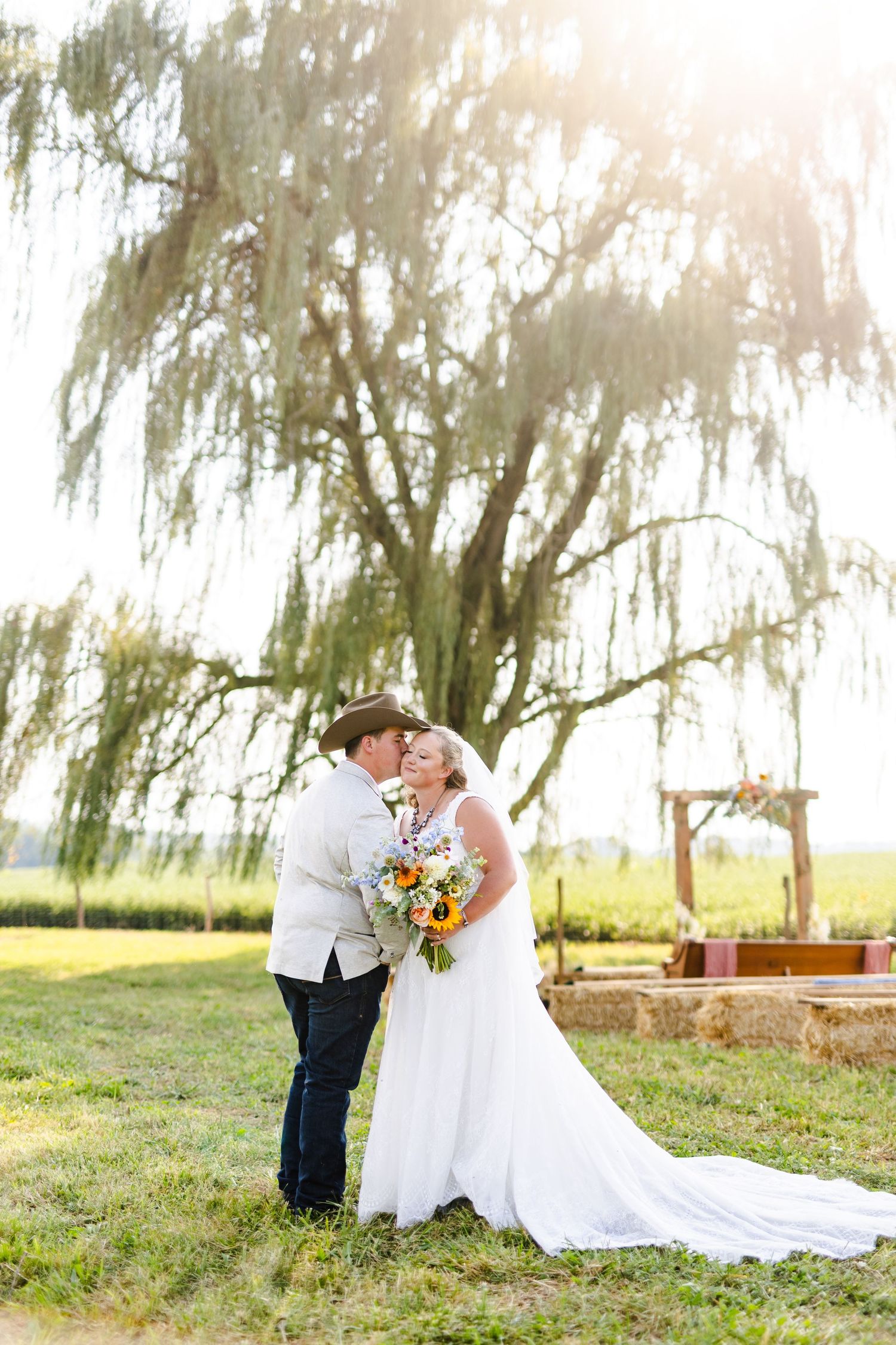 Bride and groom share a kiss under a weeping willow tree at a rustic outdoor farm wedding ceremony.