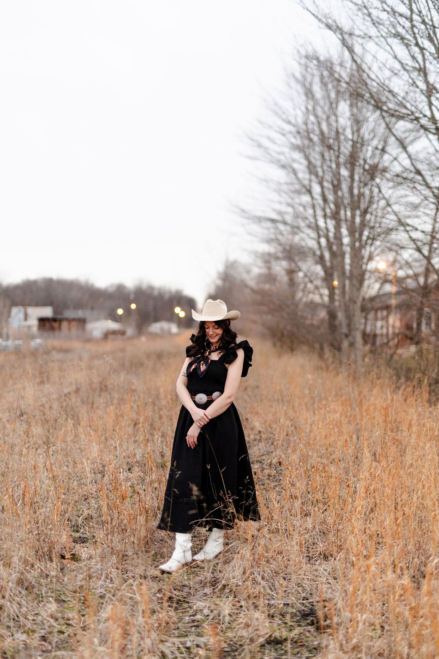 Woman in black dress and white cowboy hat standing in golden field during winter twilight.