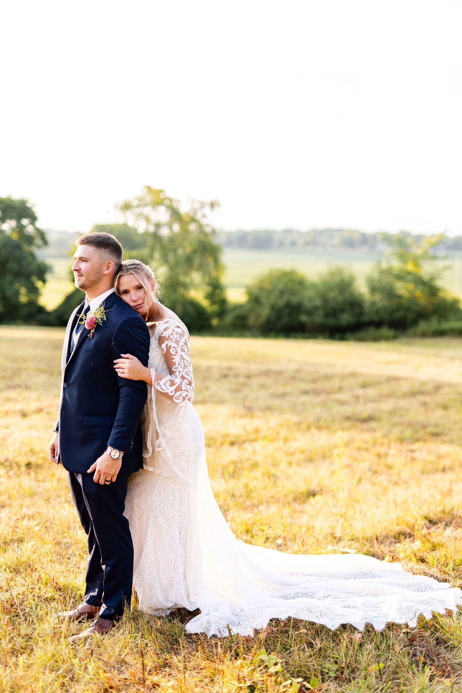 Bride leans against groom back-to-back in a sunlit field, her lace gown train flowing across the grass.