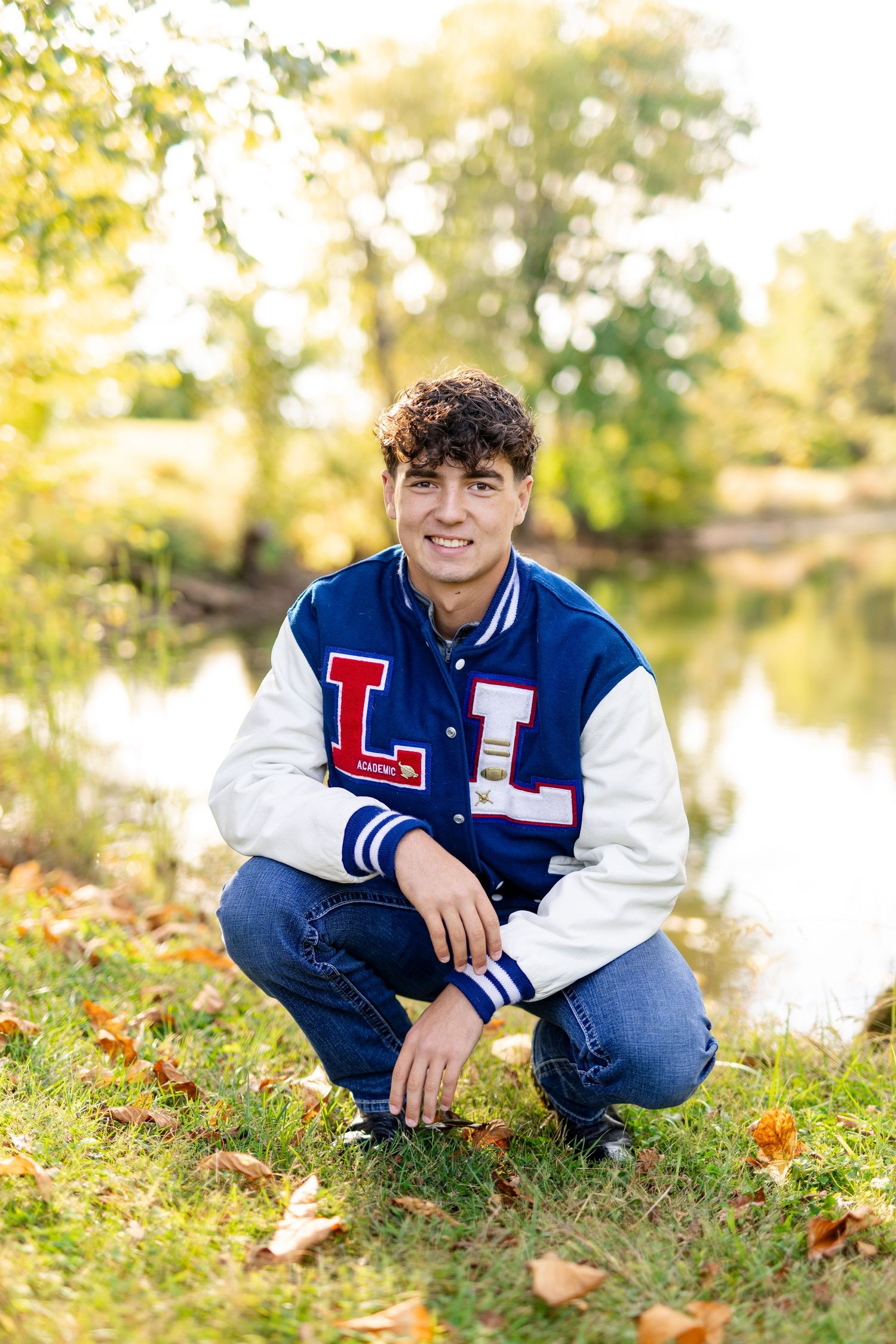 Teen boy in letterman jacket crouching by a pond surrounded by fall foliage for senior photos.