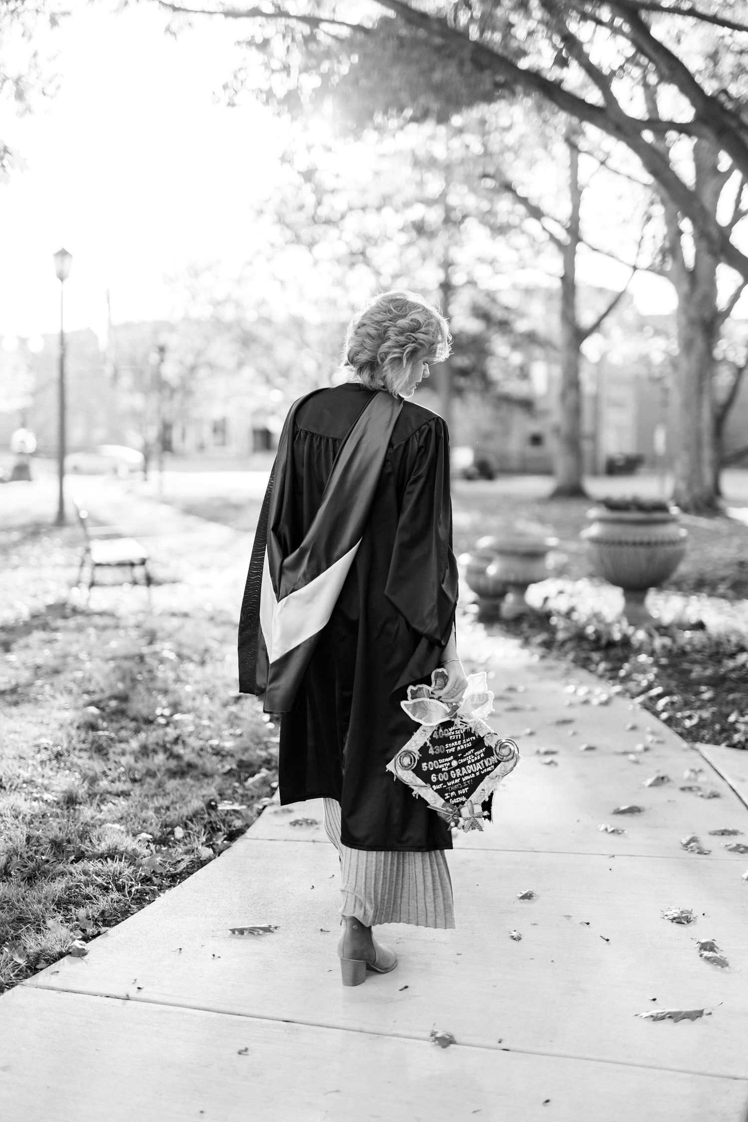 Black and white photo of graduate in cap and gown walking along sunny campus sidewalk.