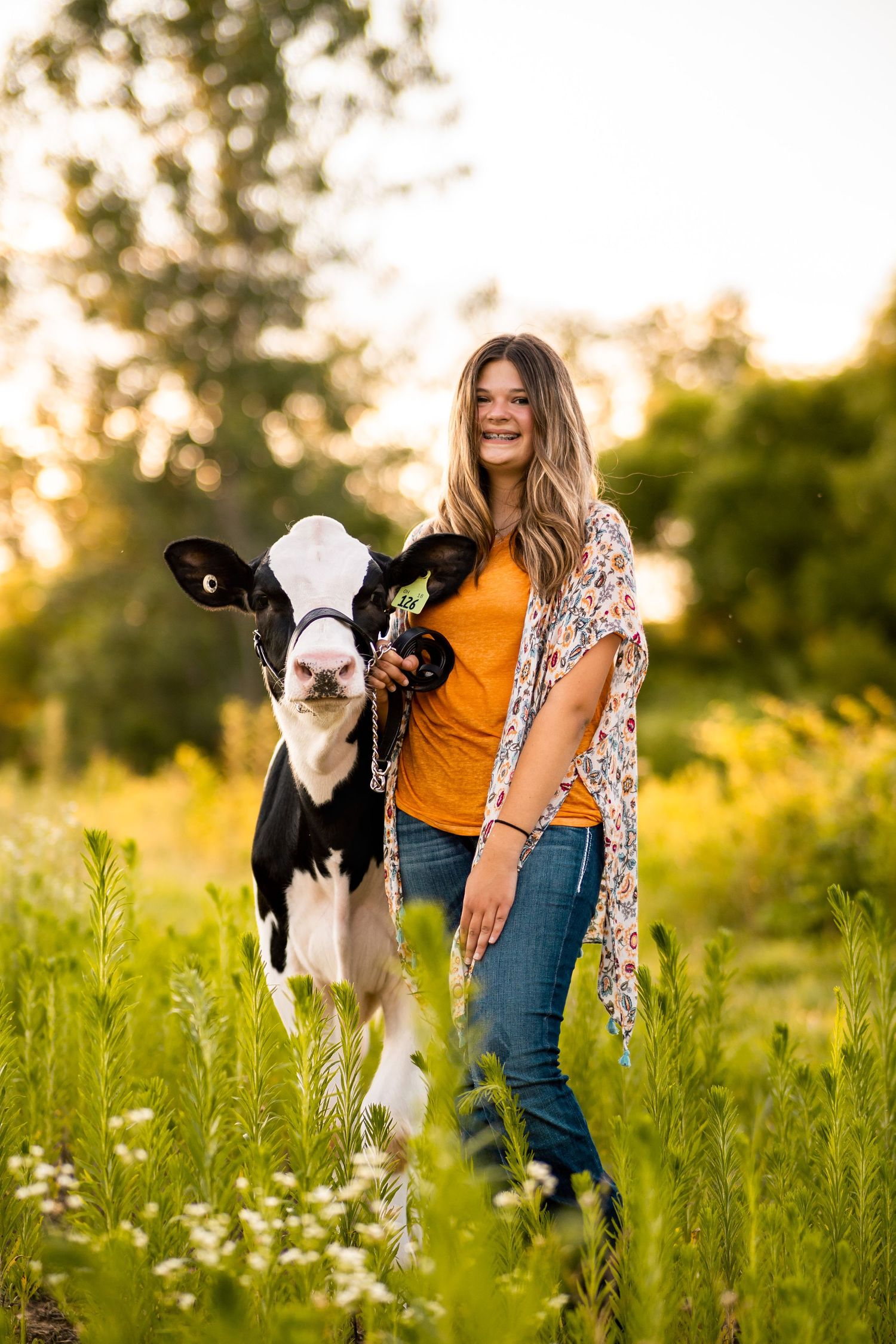 Young woman in orange shirt and jeans stands with black and white calf in sunny green meadow.