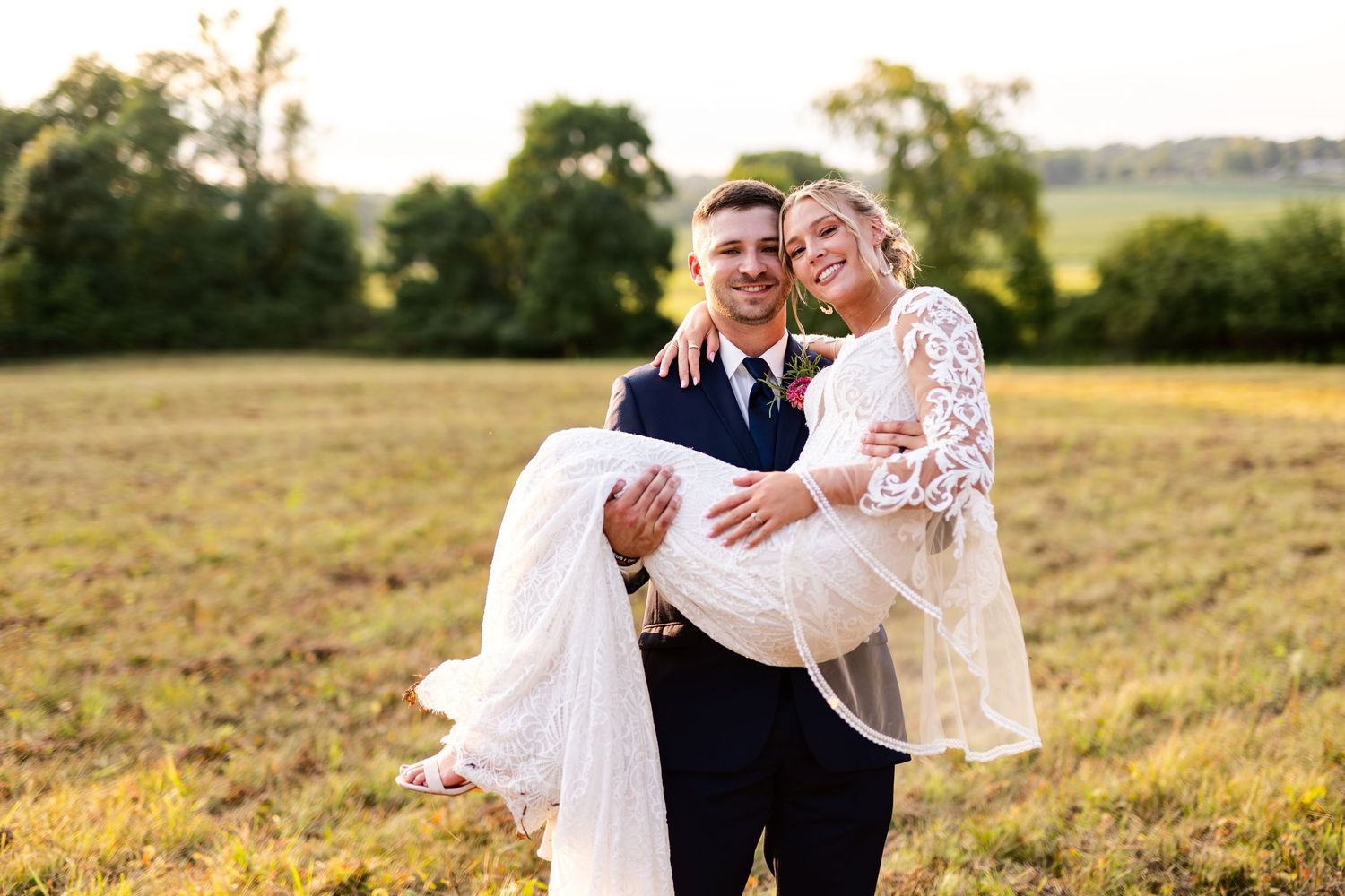 Groom in navy suit carries smiling bride in white lace dress across a sunny open field.