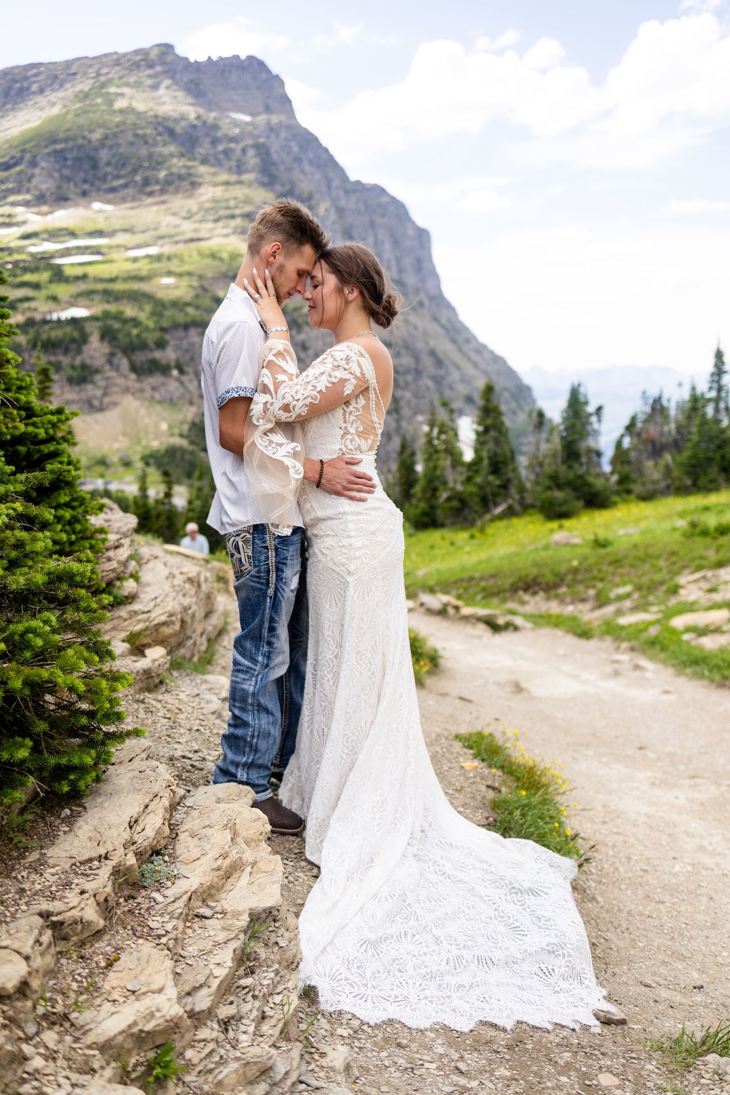 Bride and groom embrace on rocky mountain trail with stunning alpine scenery behind them.