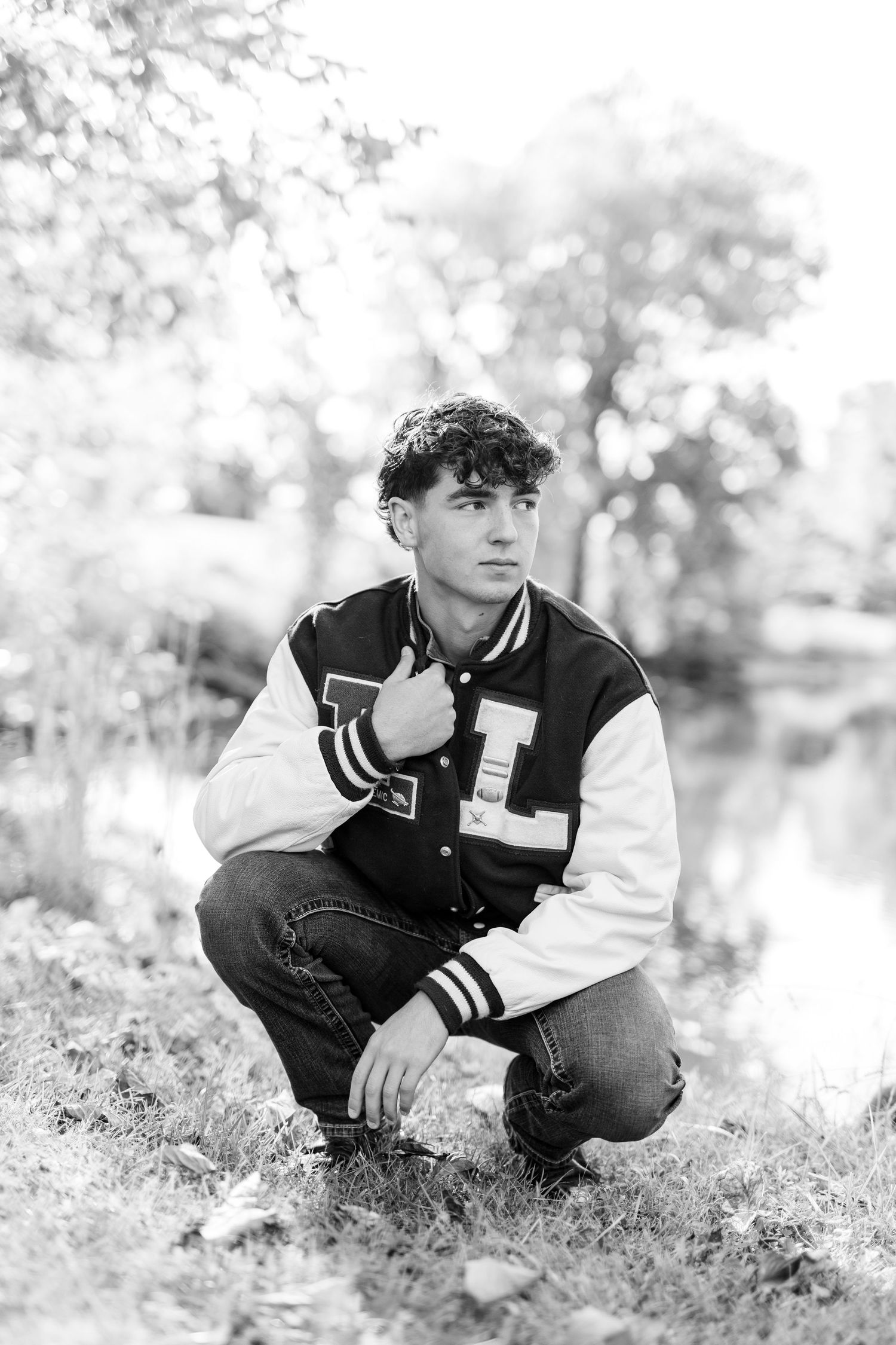Black and white photo of a young man in a varsity jacket crouching outdoors near a pond.