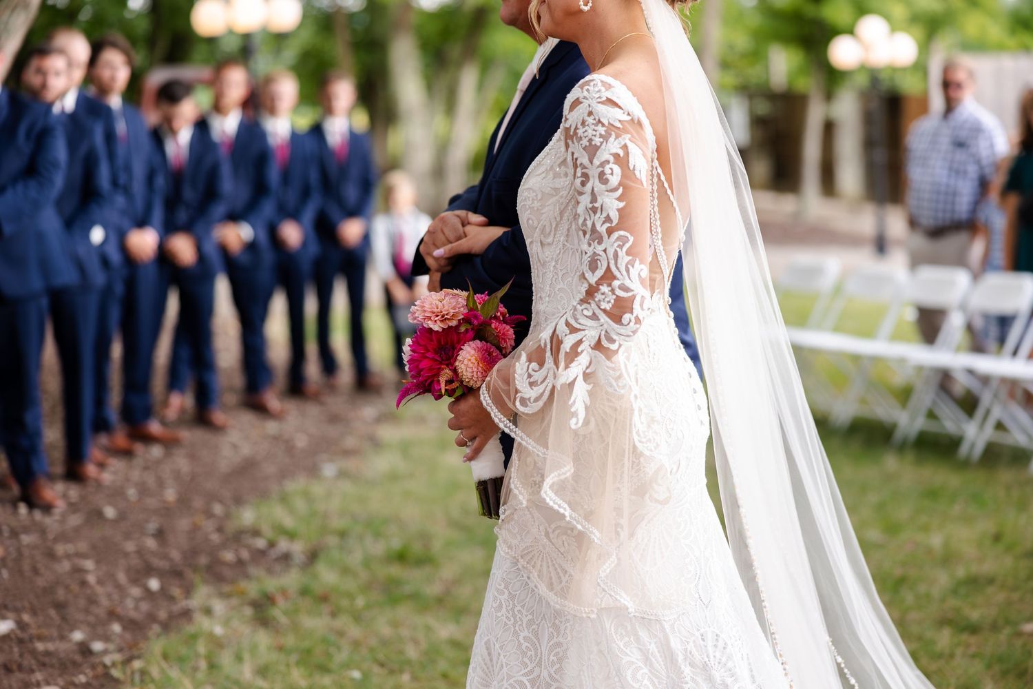 Bride in lace wedding gown with veil walks down the aisle holding a vibrant pink and red bouquet.