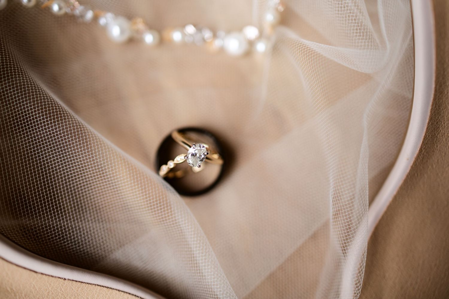 Close-up of a gold diamond engagement ring resting on a bridal veil with pearl necklace detail.
