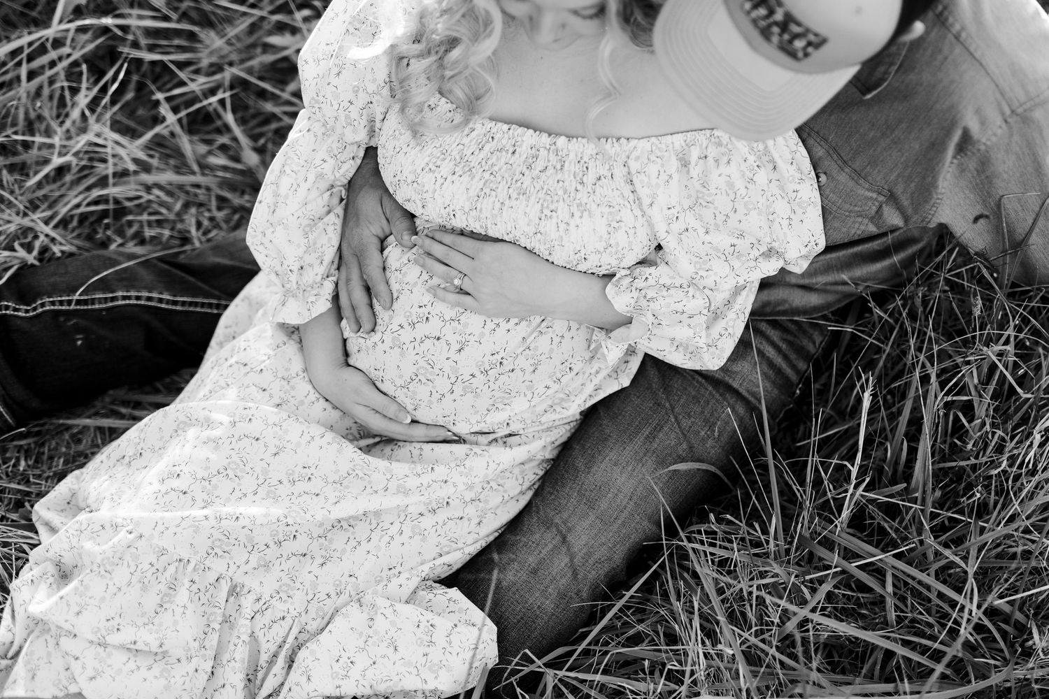 Black and white maternity photo of pregnant woman in lace dress sitting in grass with partner.