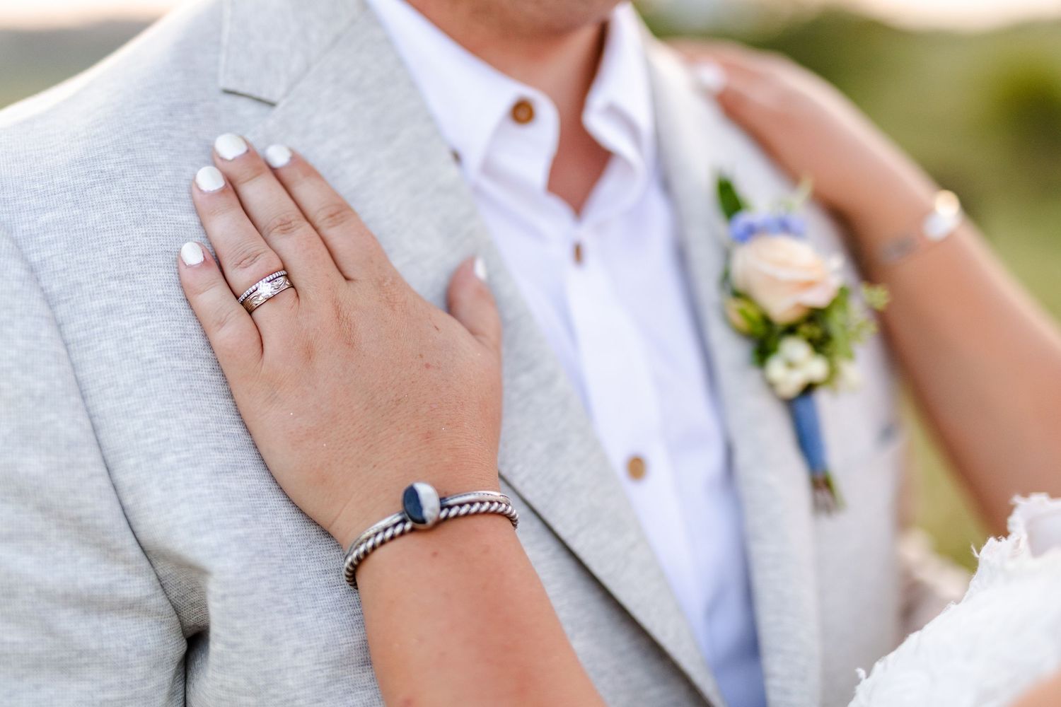 Close-up of bride's hand with white manicure, silver rings, and bracelet resting on groom's gray suit jacket.