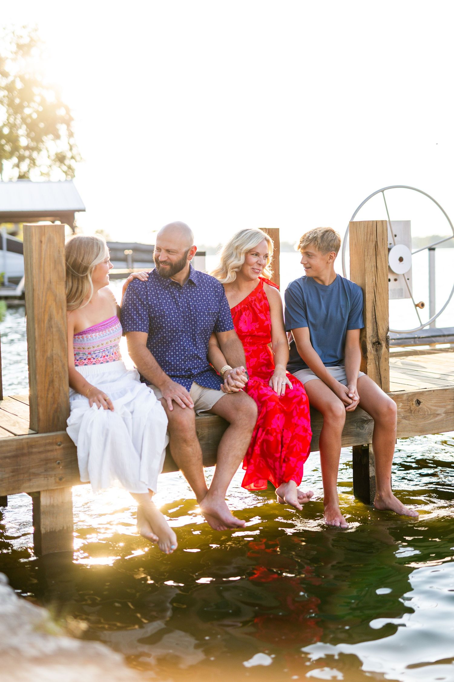 Family of four sitting on a sunny dock with feet dangling over sparkling water in summer attire.