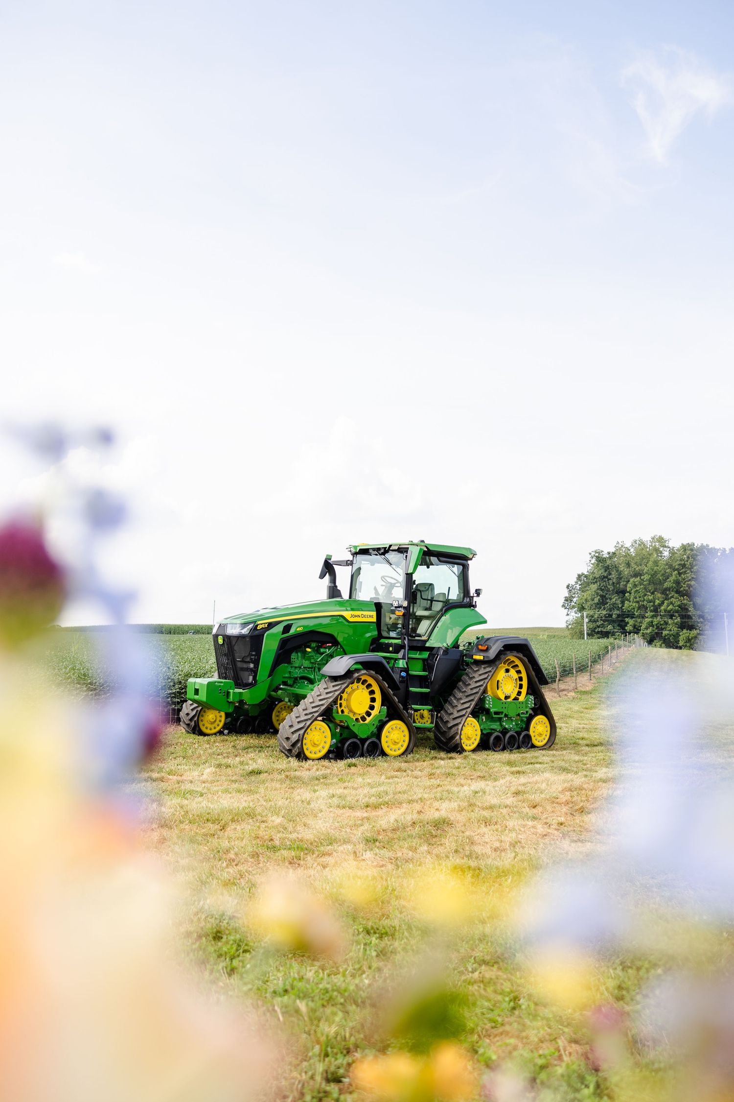Green John Deere tractor in a farm field framed by colorful blurred wildflowers in the foreground.