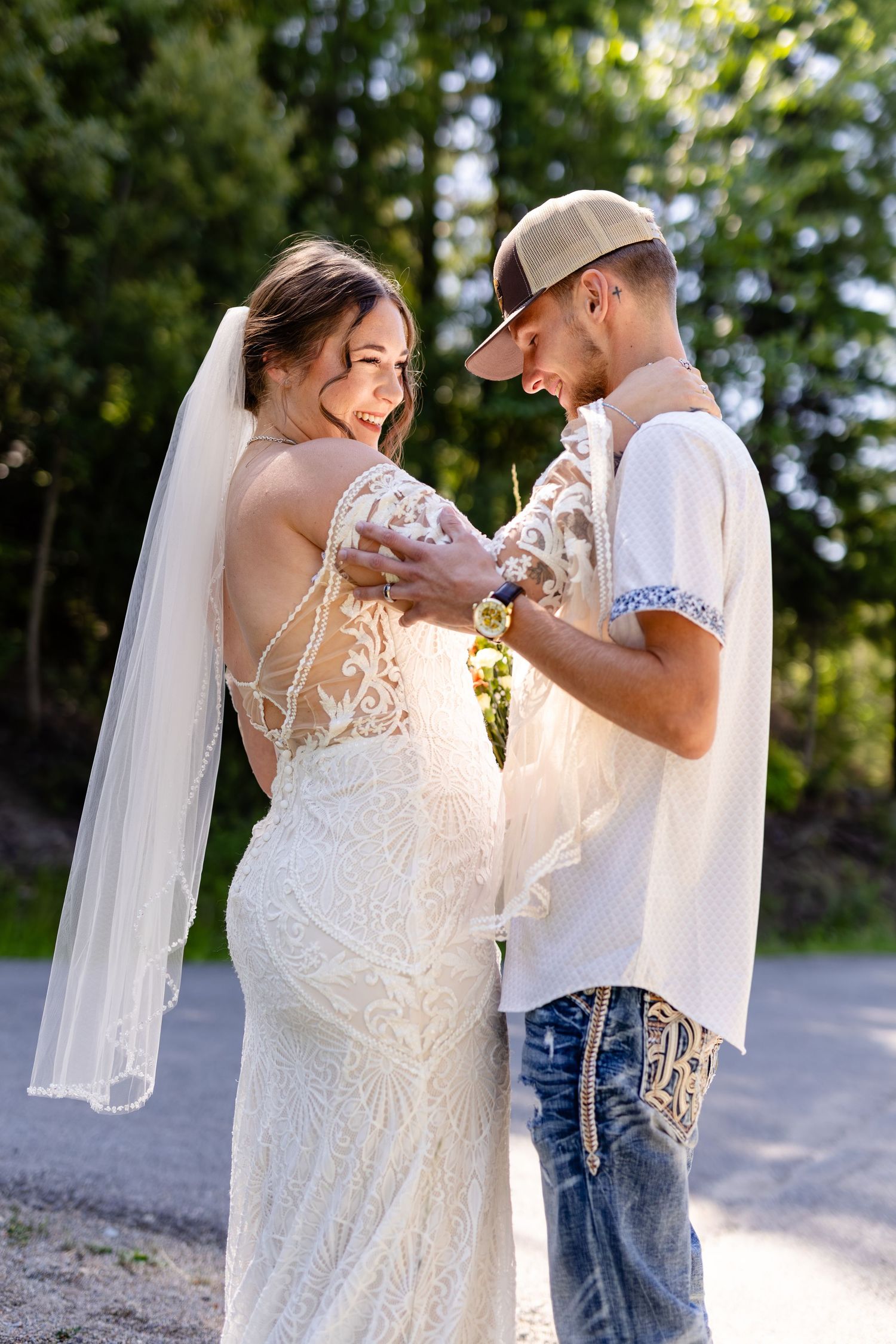 Groom in white shirt and jeans embraces smiling bride in lace wedding dress outdoors.