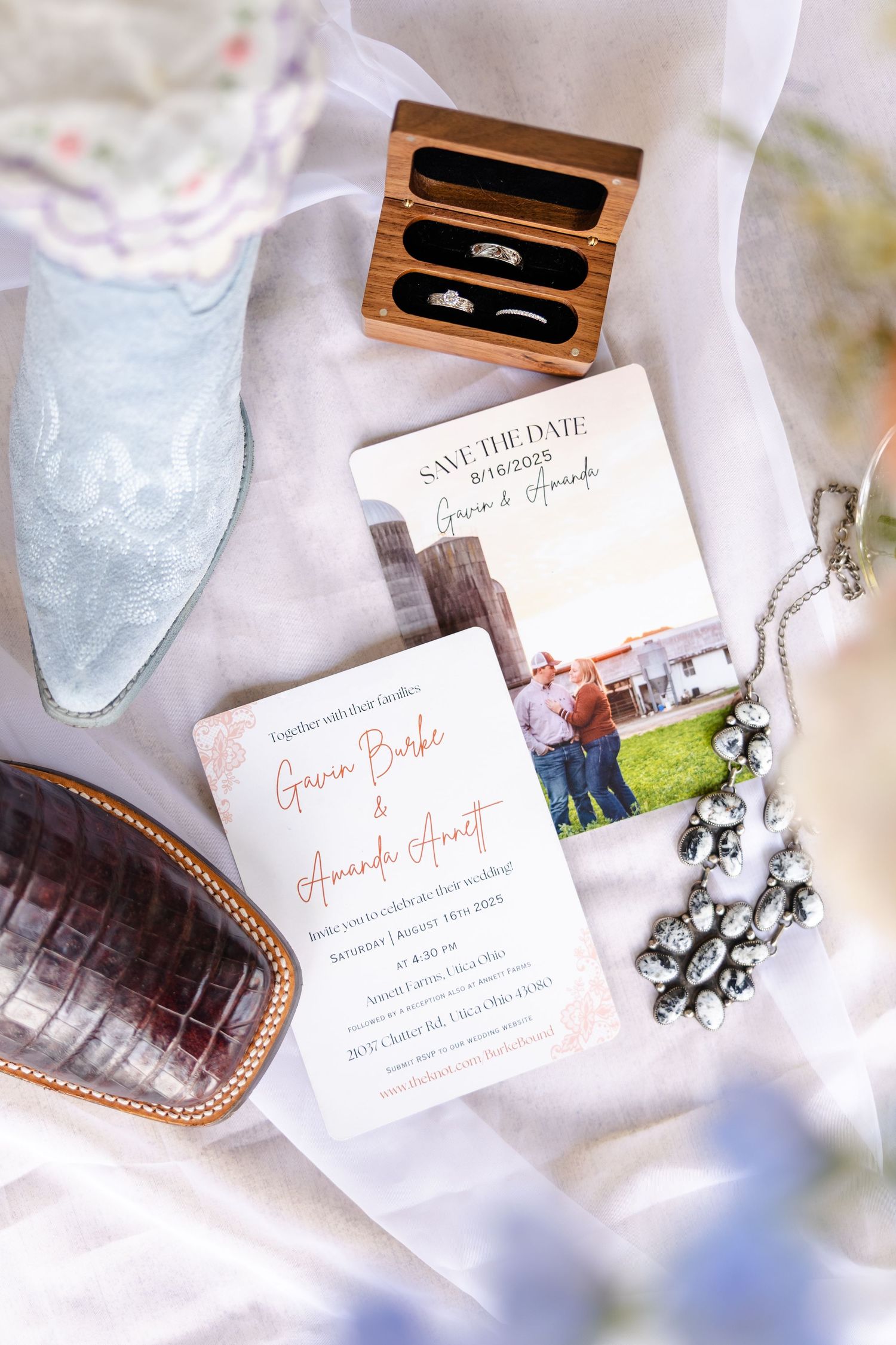 Rustic wedding flat lay with cowboy boot, floral invitations, rings in wooden box, and crystal necklace on white fabric.