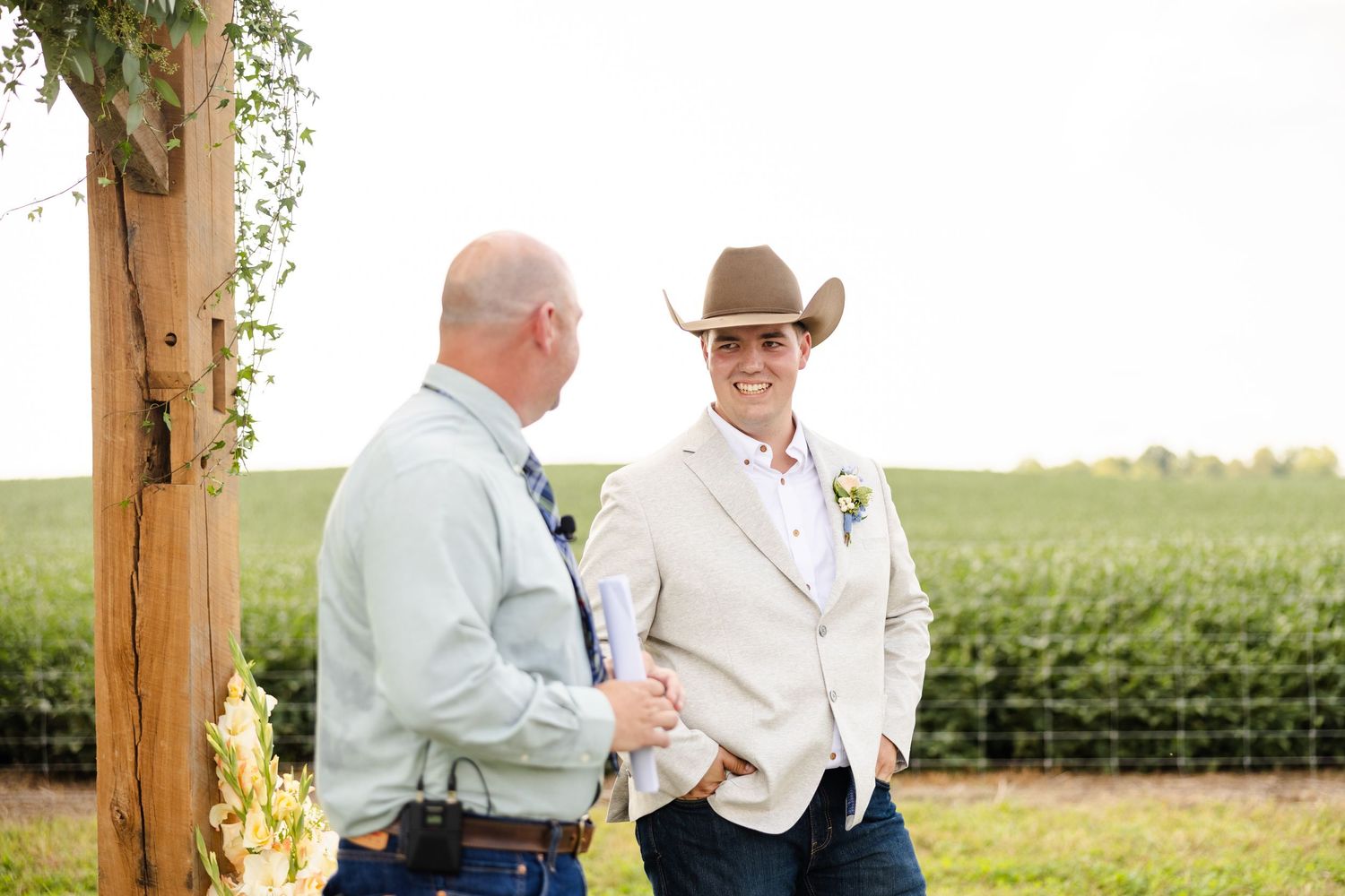 Groom in cowboy hat and tan blazer laughing with a guest near a wooden arch at outdoor wedding.