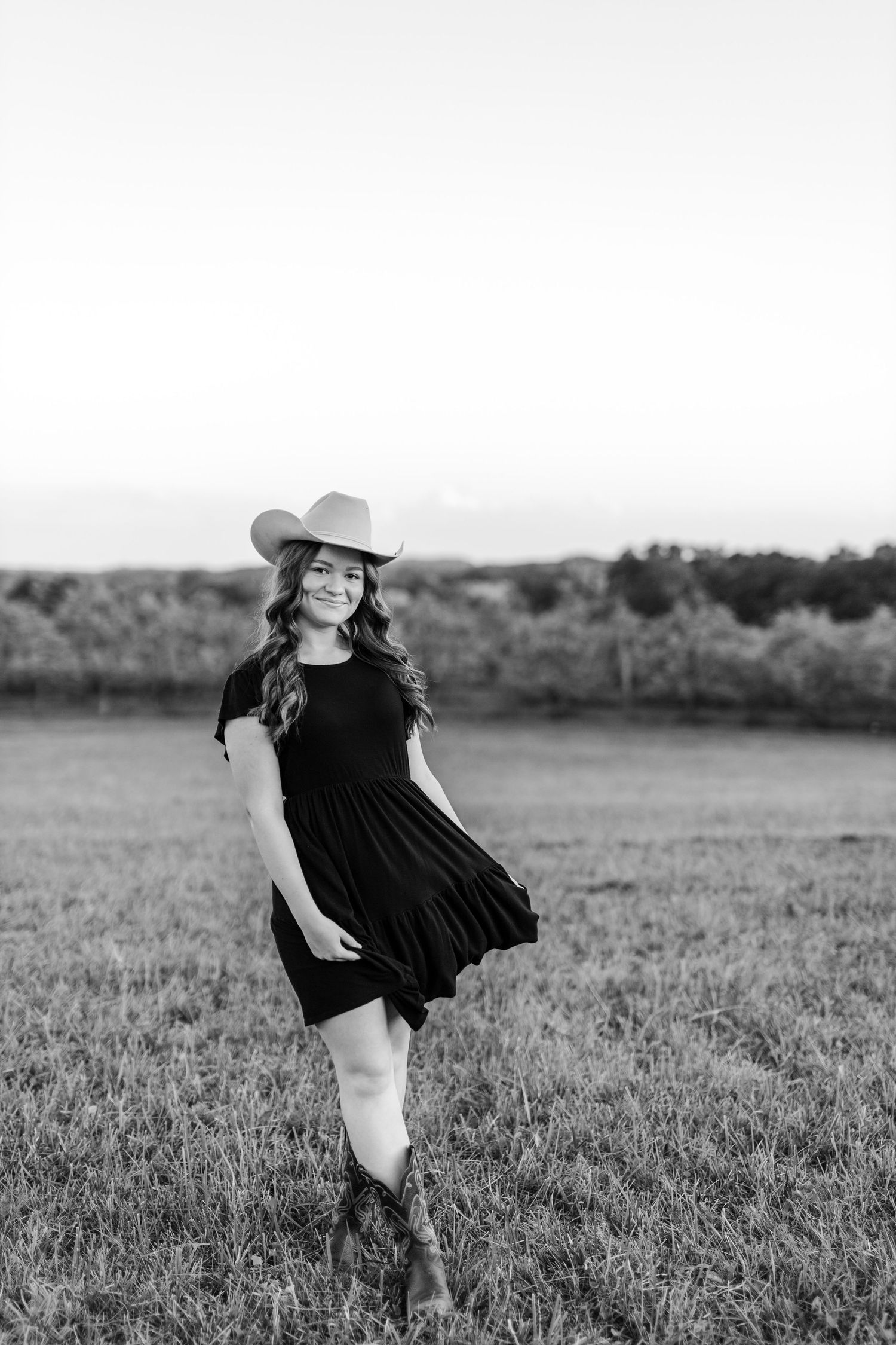 Black and white photo of a young woman in a cowboy hat and dress posing in a field at sunset.