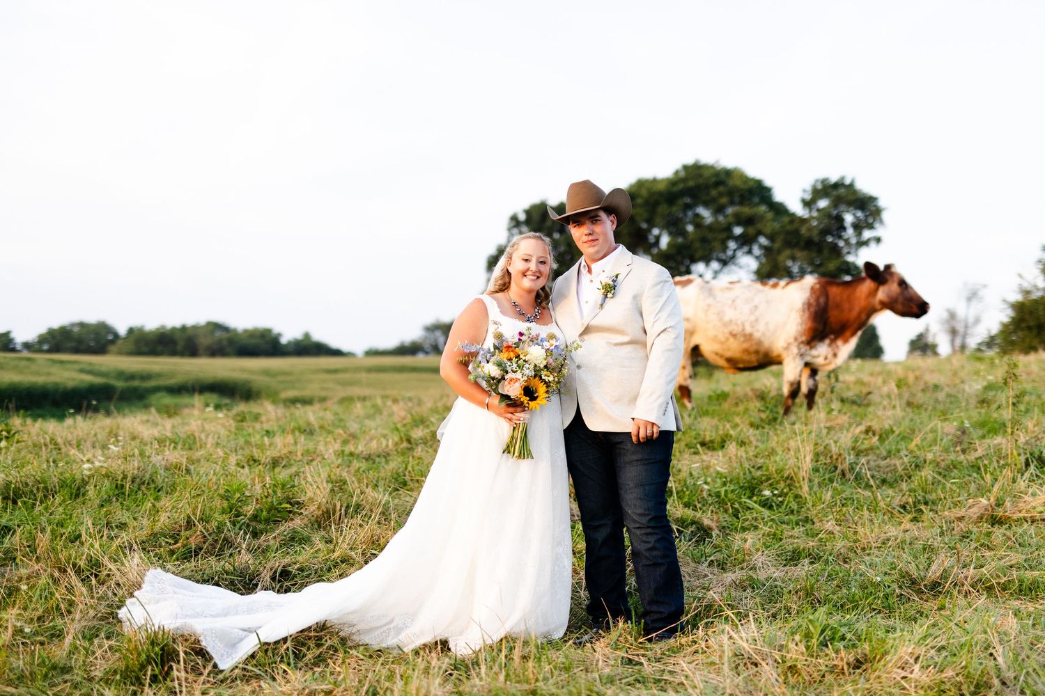 Smiling bride and groom pose with a shorthorn cow in a rustic farm field at their country wedding.