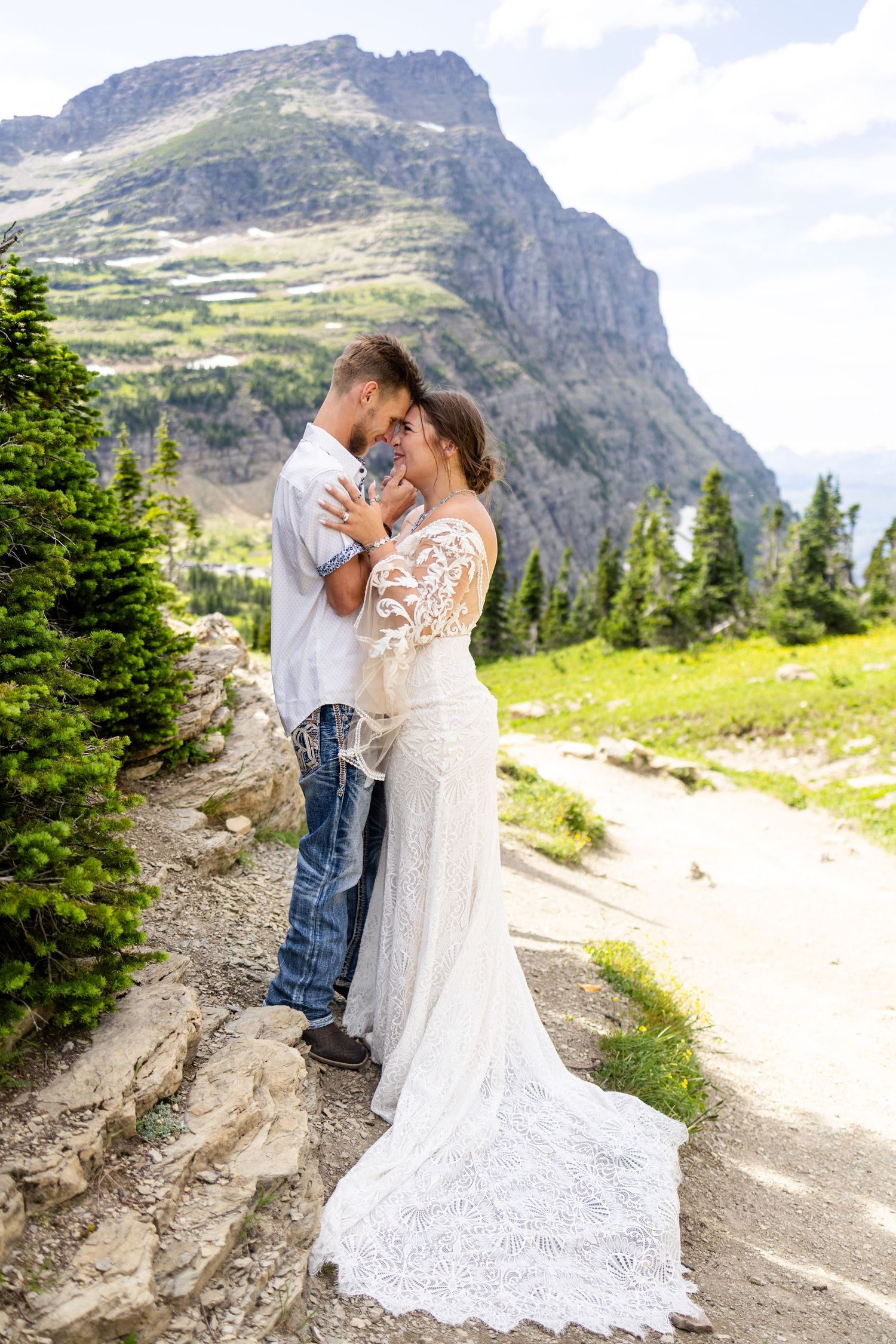 Bride and groom embrace on rocky mountain path with scenic peaks and evergreen trees in background.