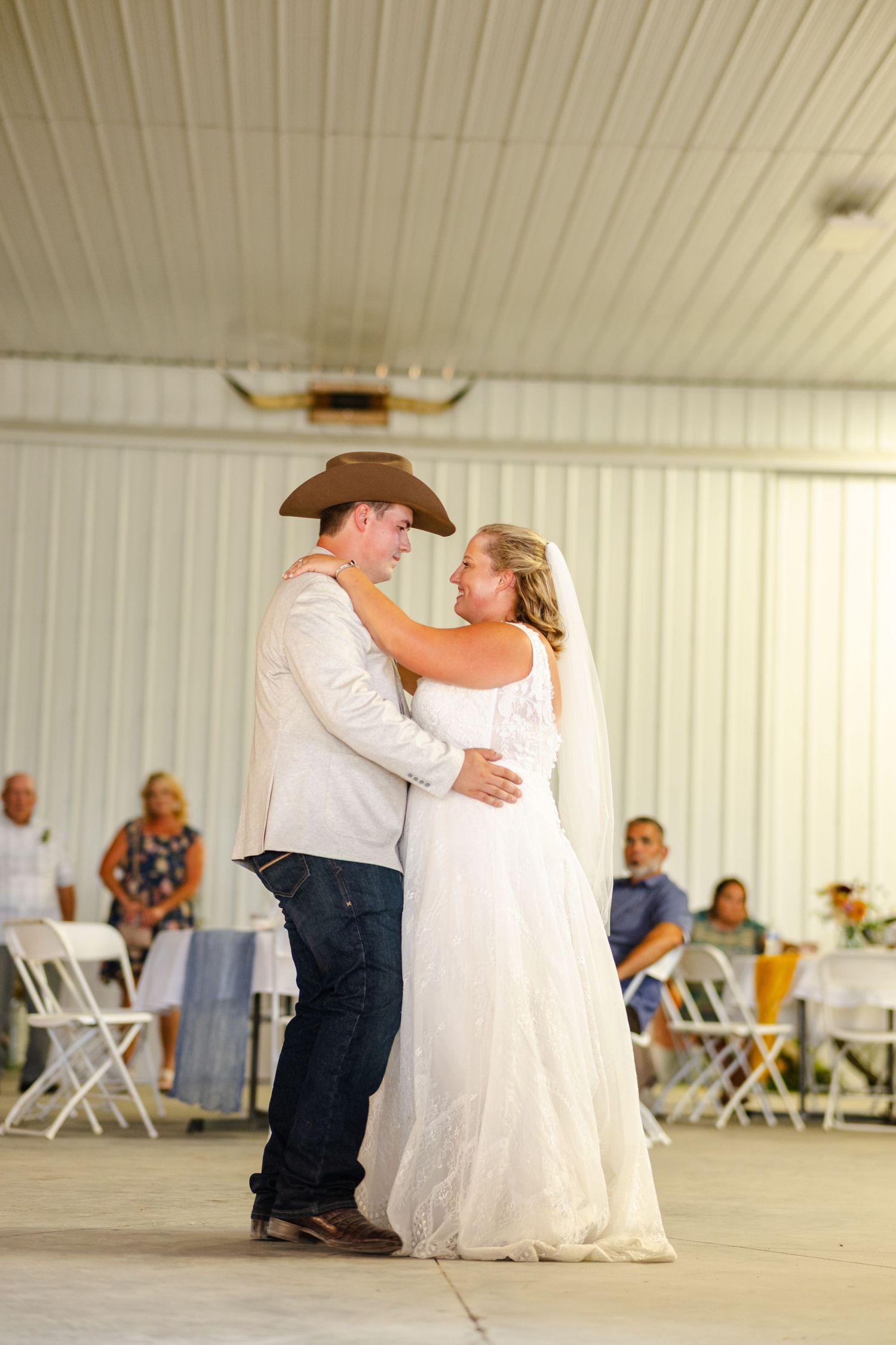 Bride and groom share their first dance in a white barn venue while guests watch from the background.