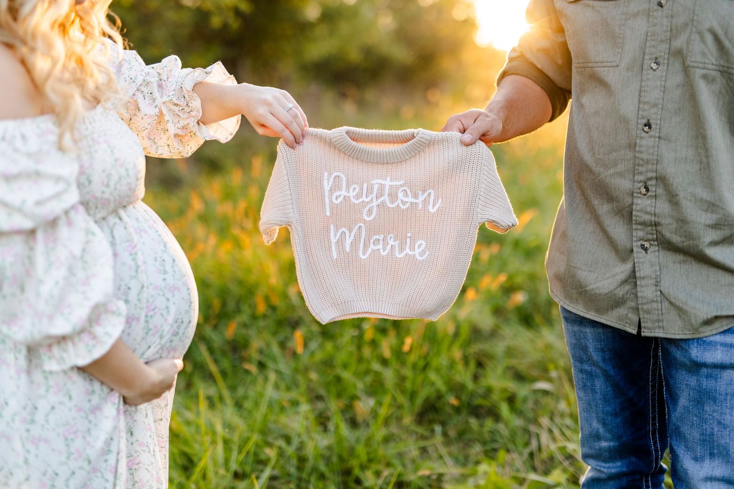 Expecting couple holding pink baby onesie reading Peyton Marie during golden hour maternity photo session.