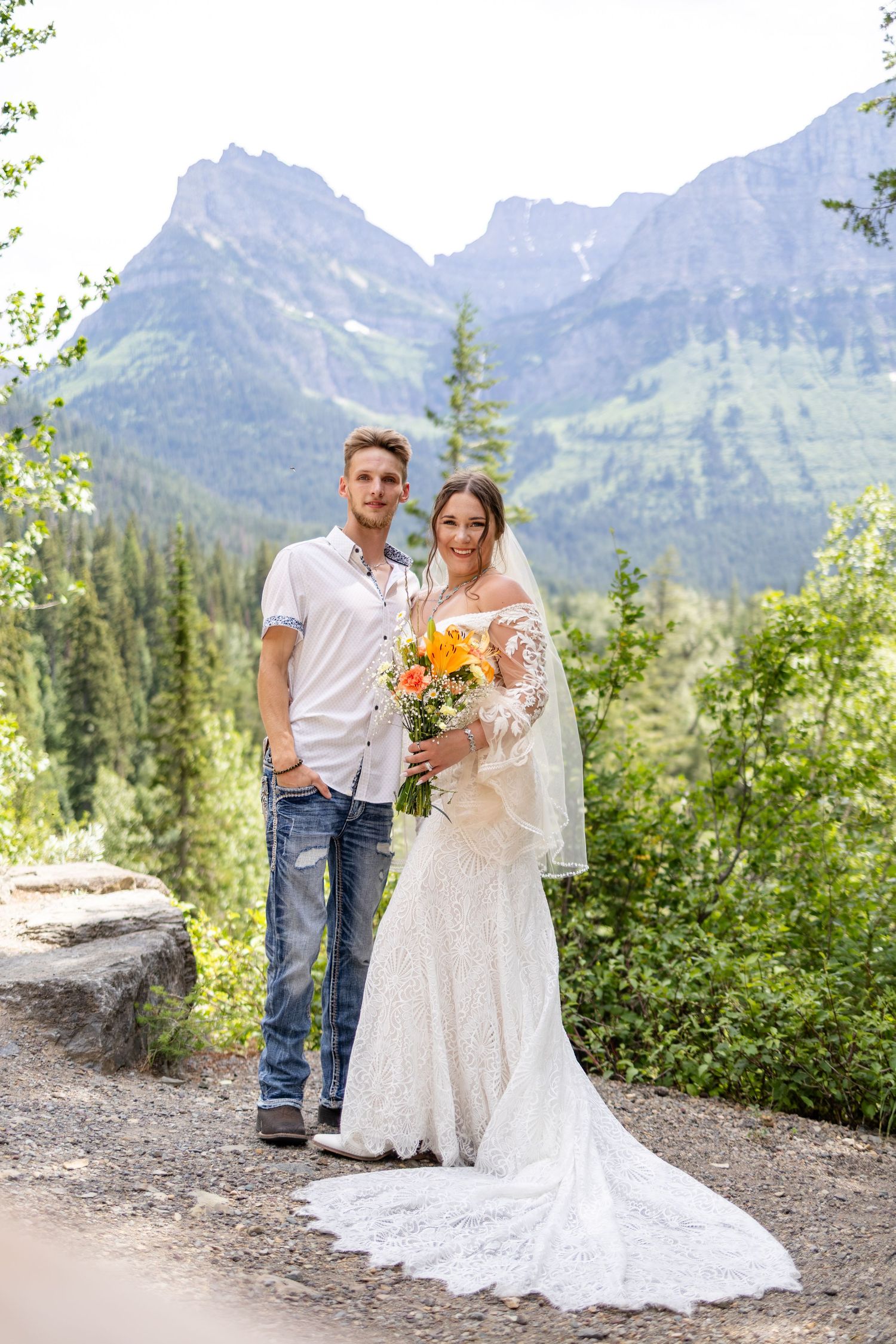 Couple poses together on mountain trail, bride in lace gown holding orange bouquet with stunning rocky peaks behind them.