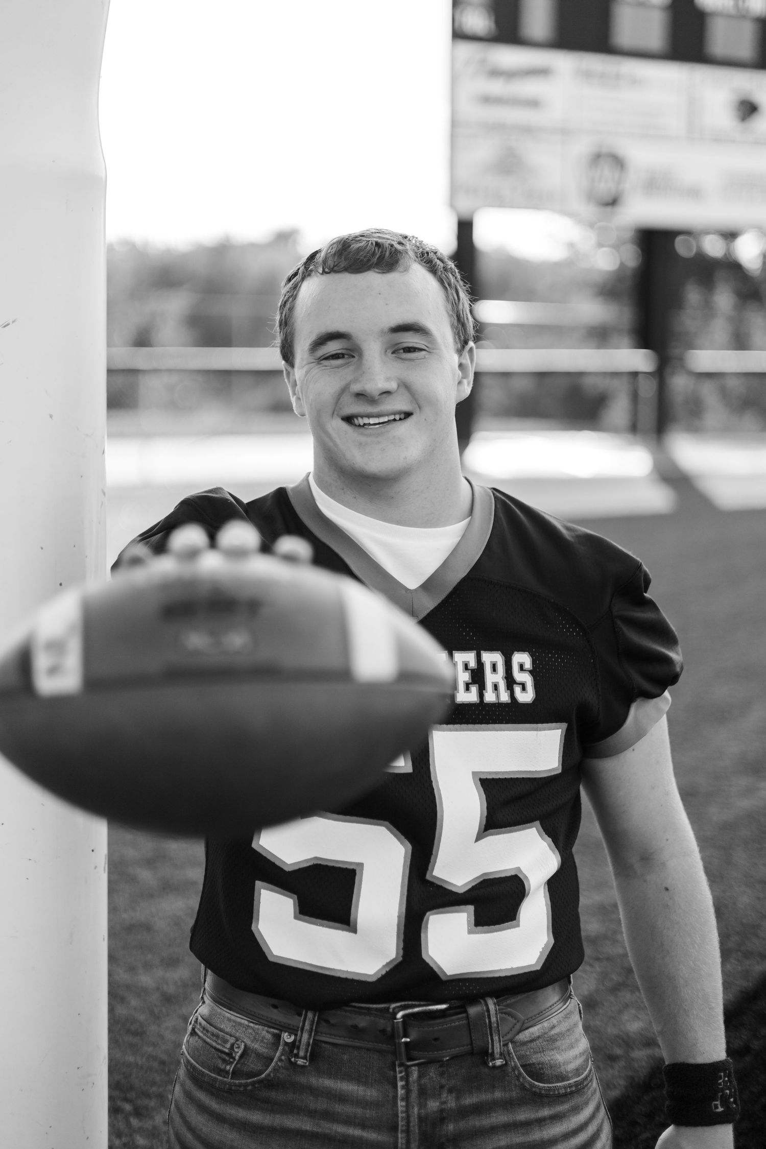 Black and white photo of a smiling teen in football jersey number 55 holding a football at a stadium.
