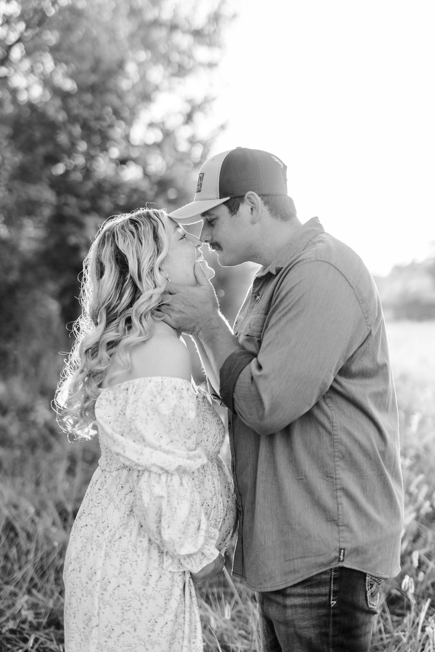 Black and white photo of a couple sharing a romantic almost-kiss outdoors, man in cap and shirt, woman in floral dress.