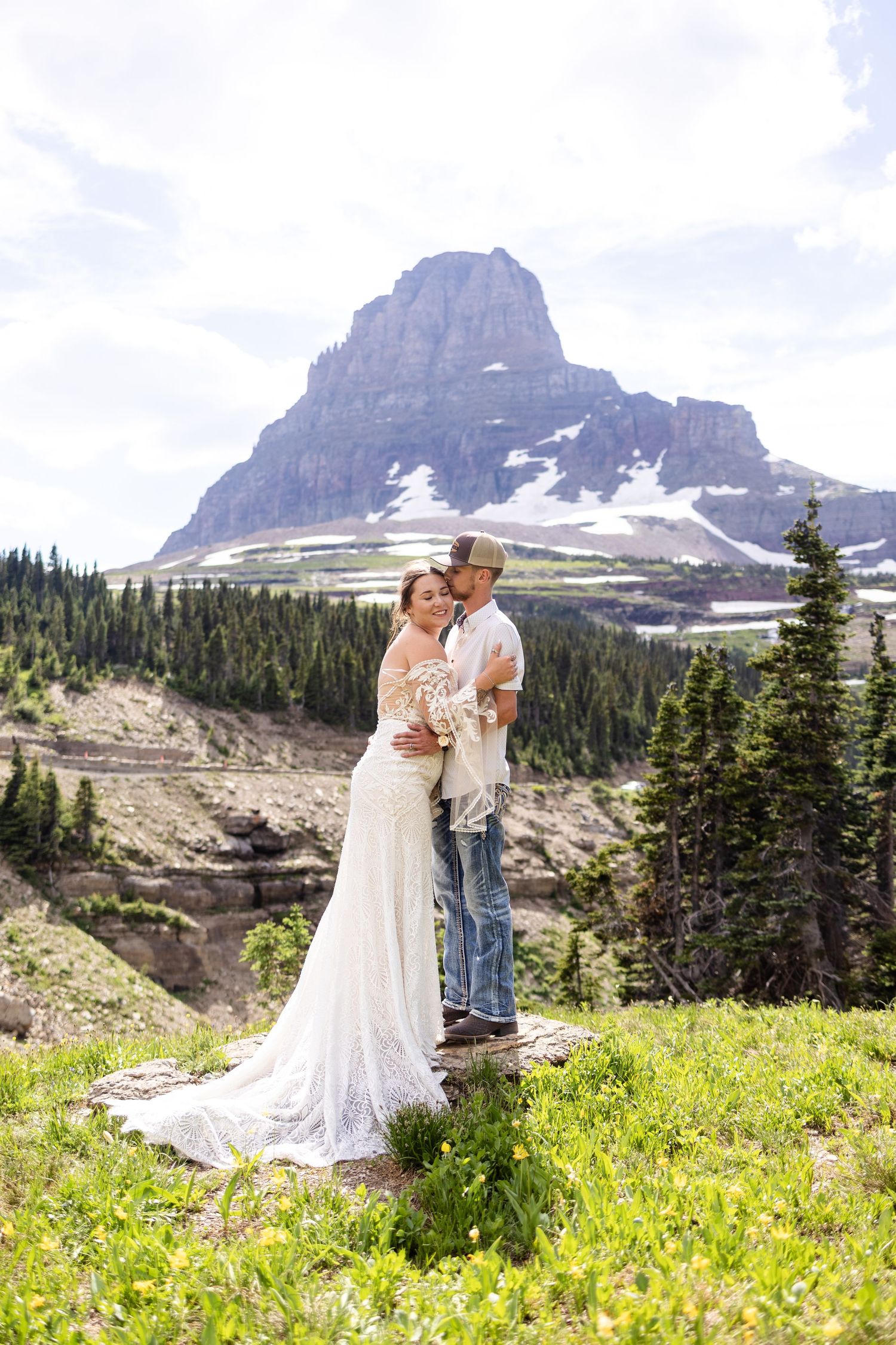 Bride and groom embracing on mountain meadow with snow-capped peak and evergreen forest behind them.