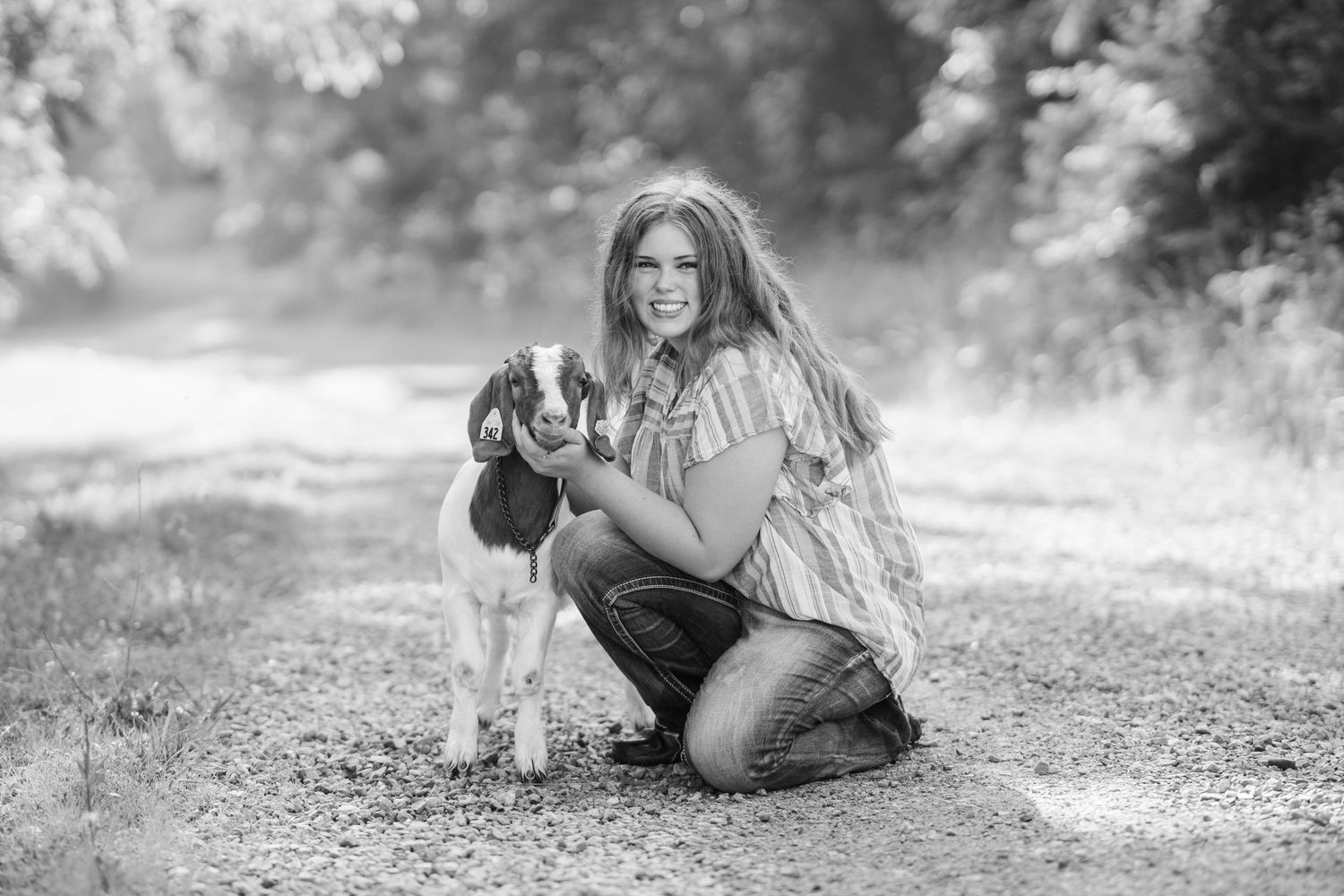 Young woman kneeling on a dirt path holding a small goat, smiling in a black and white outdoor photo.