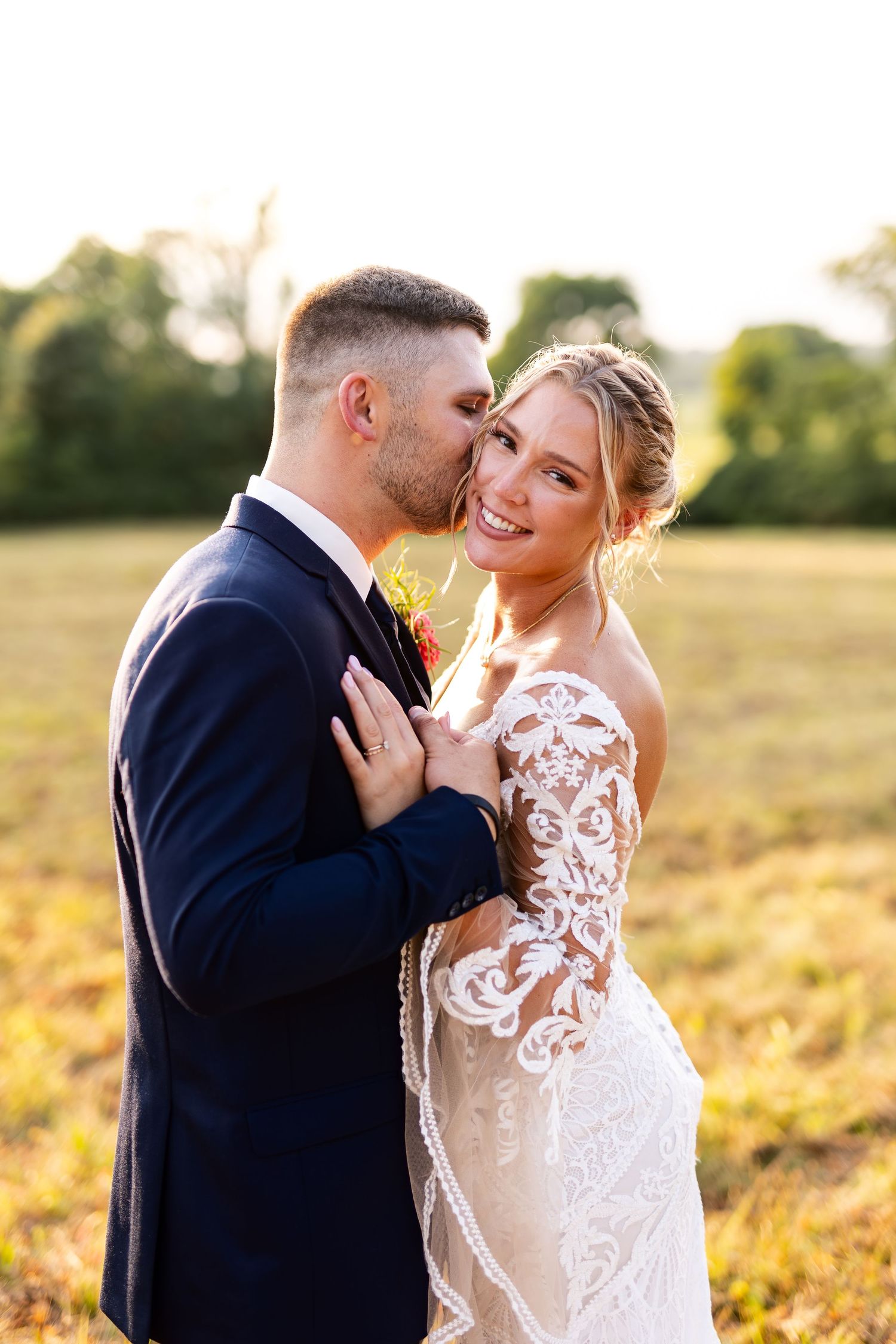 Groom kisses bride on cheek as she smiles in a lace wedding gown at golden hour.