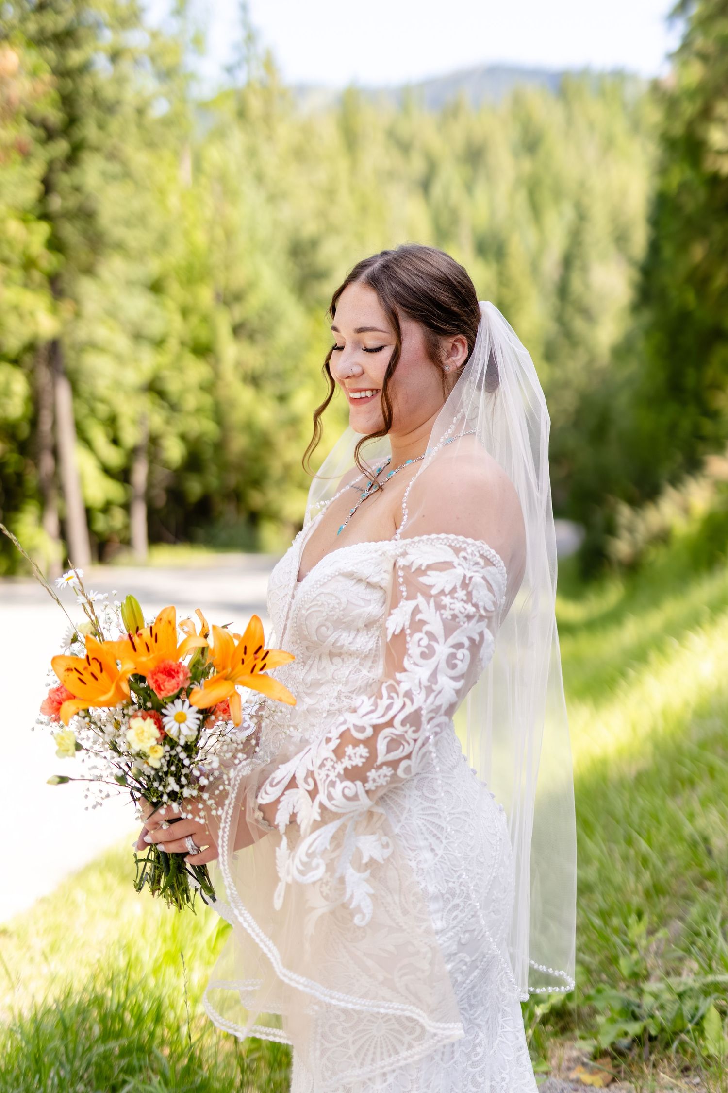Bride in off-shoulder lace wedding dress smiles while holding vibrant orange lily bouquet outdoors.