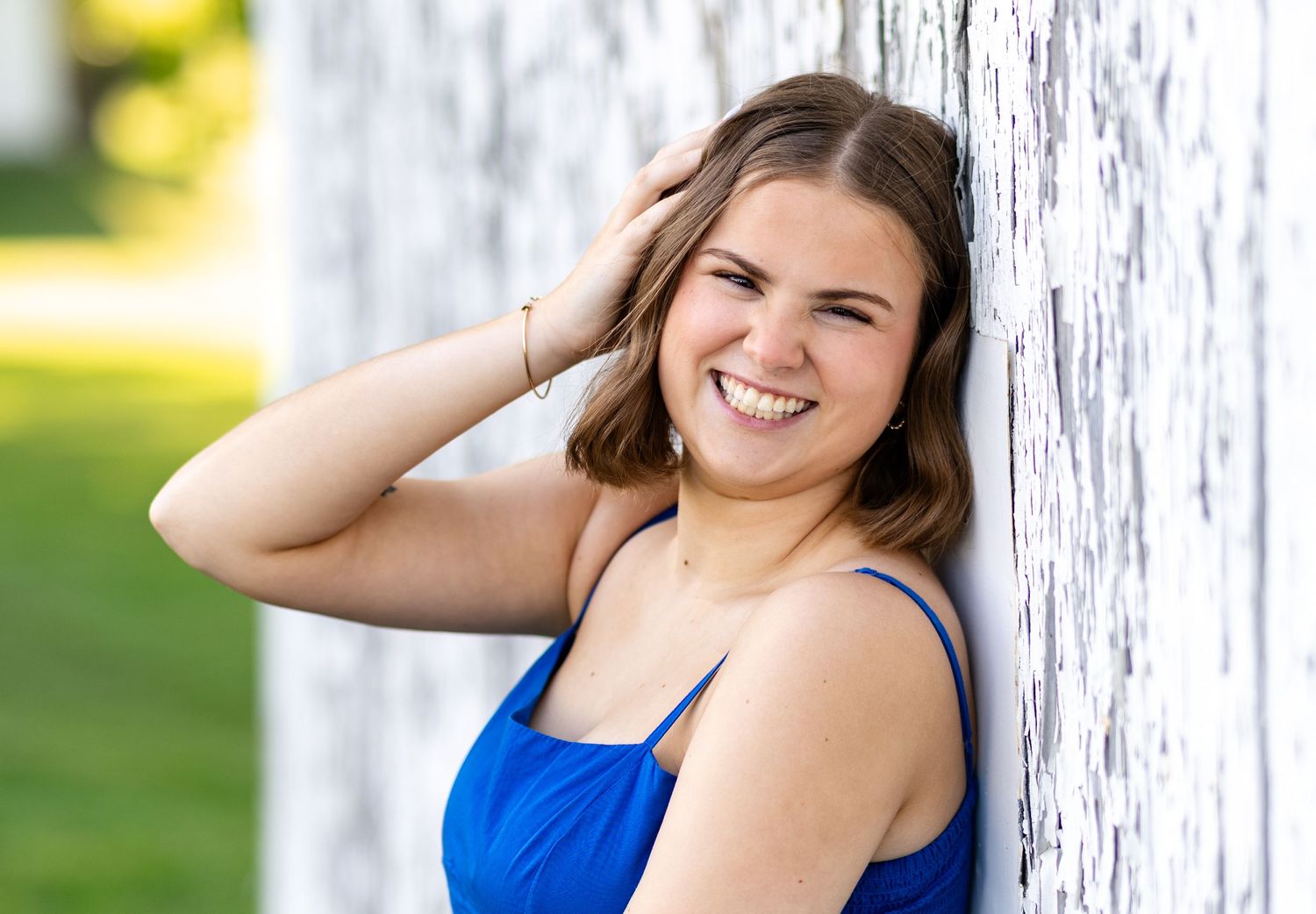 Smiling young woman in blue dress leaning against white wooden wall outdoors.