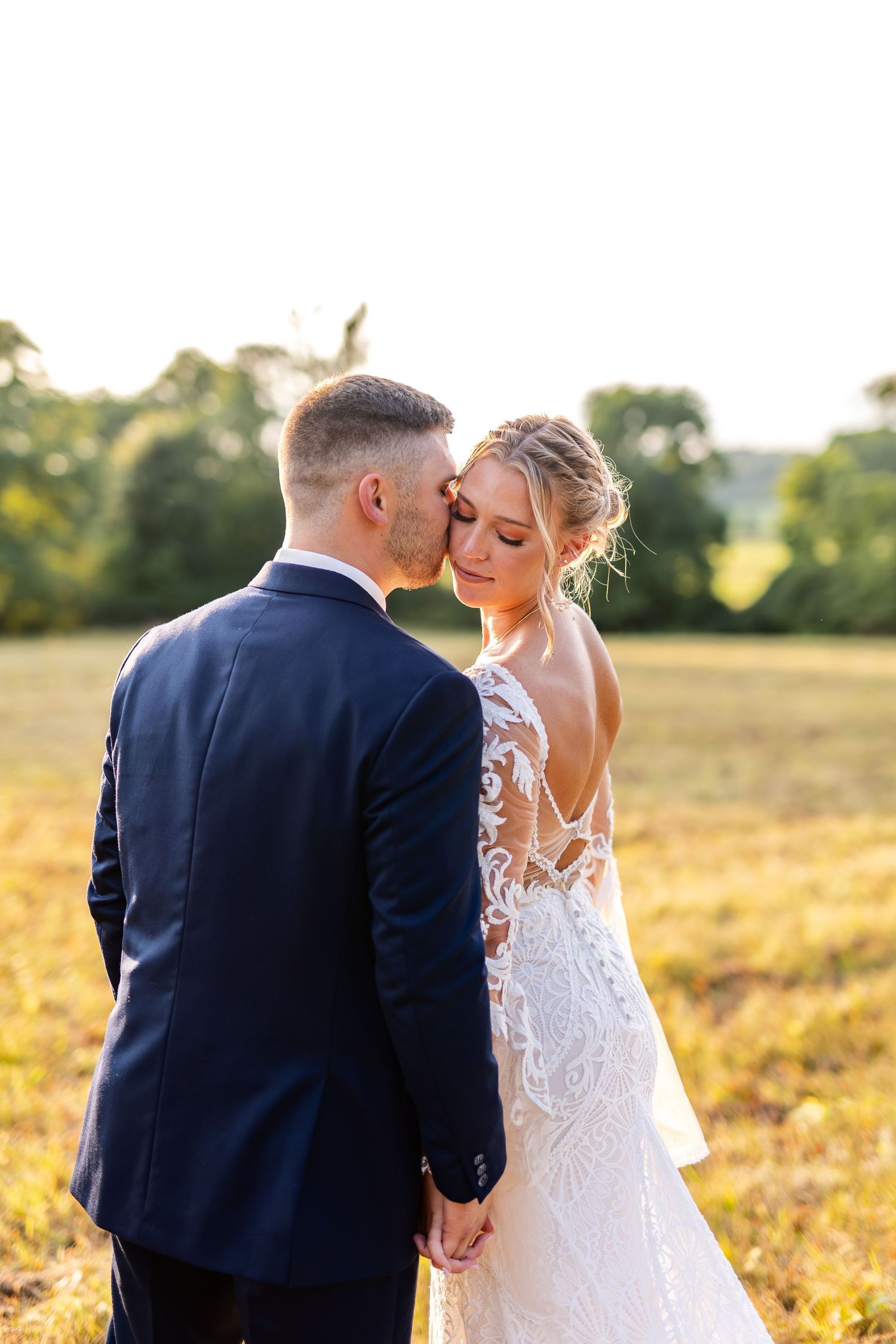 Groom tenderly kisses bride on the temple during golden hour portrait in an open countryside field.