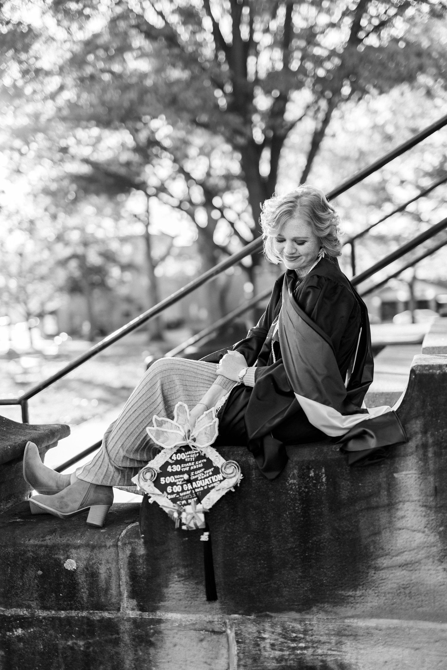 Graduate in cap and gown sitting on outdoor steps holding diploma in black and white photo.
