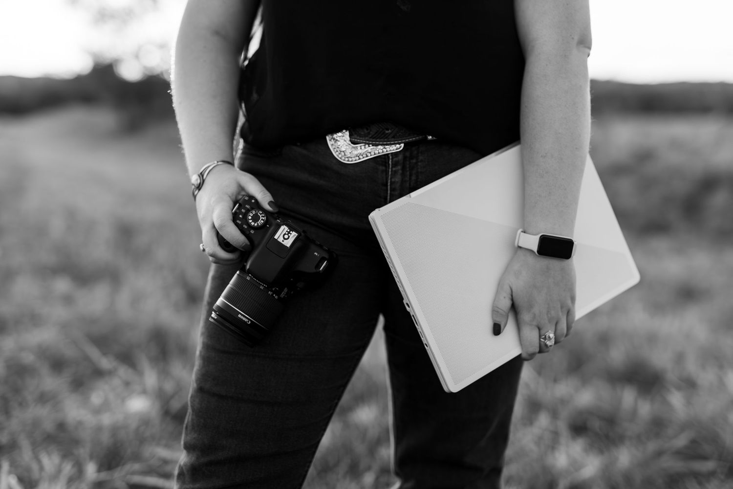 Black and white photo of Laura holding a camera and laptop in an outdoor field setting.