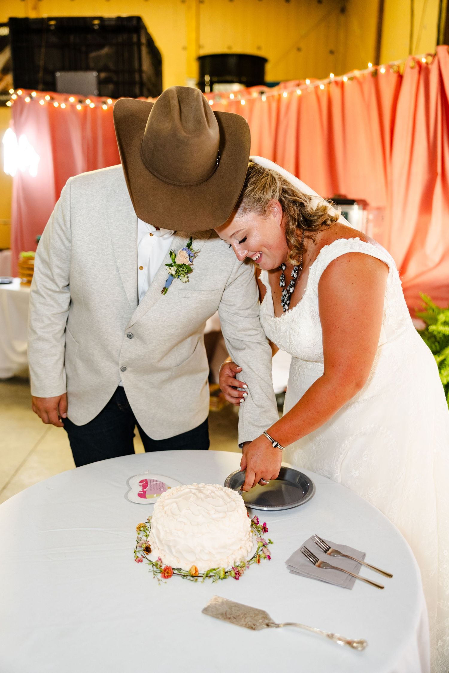 Bride and groom laugh together while cutting their white wedding cake decorated with colorful flowers.
