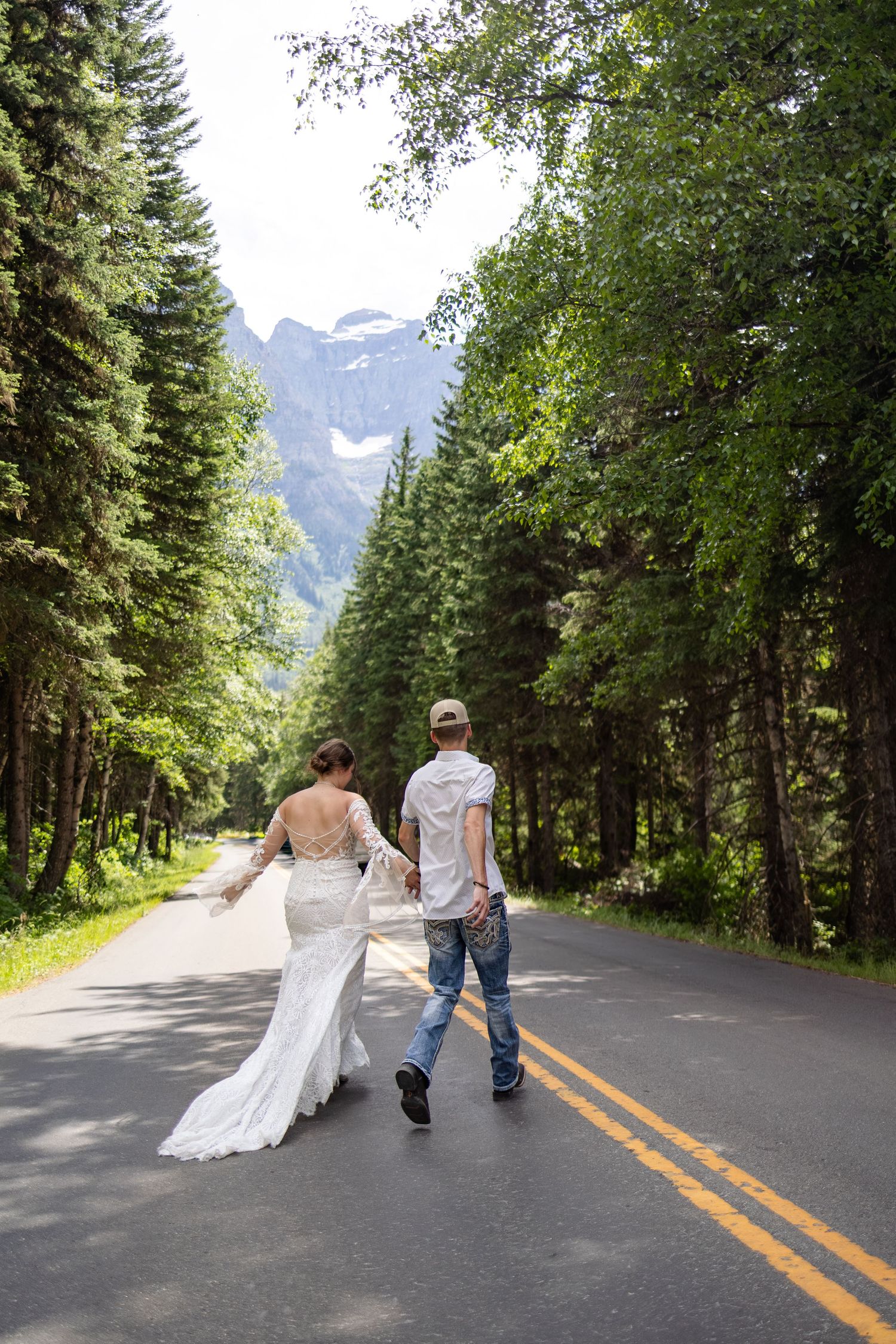Bride and groom walk hand-in-hand down scenic tree-lined mountain road with snow-capped peaks in the distance.