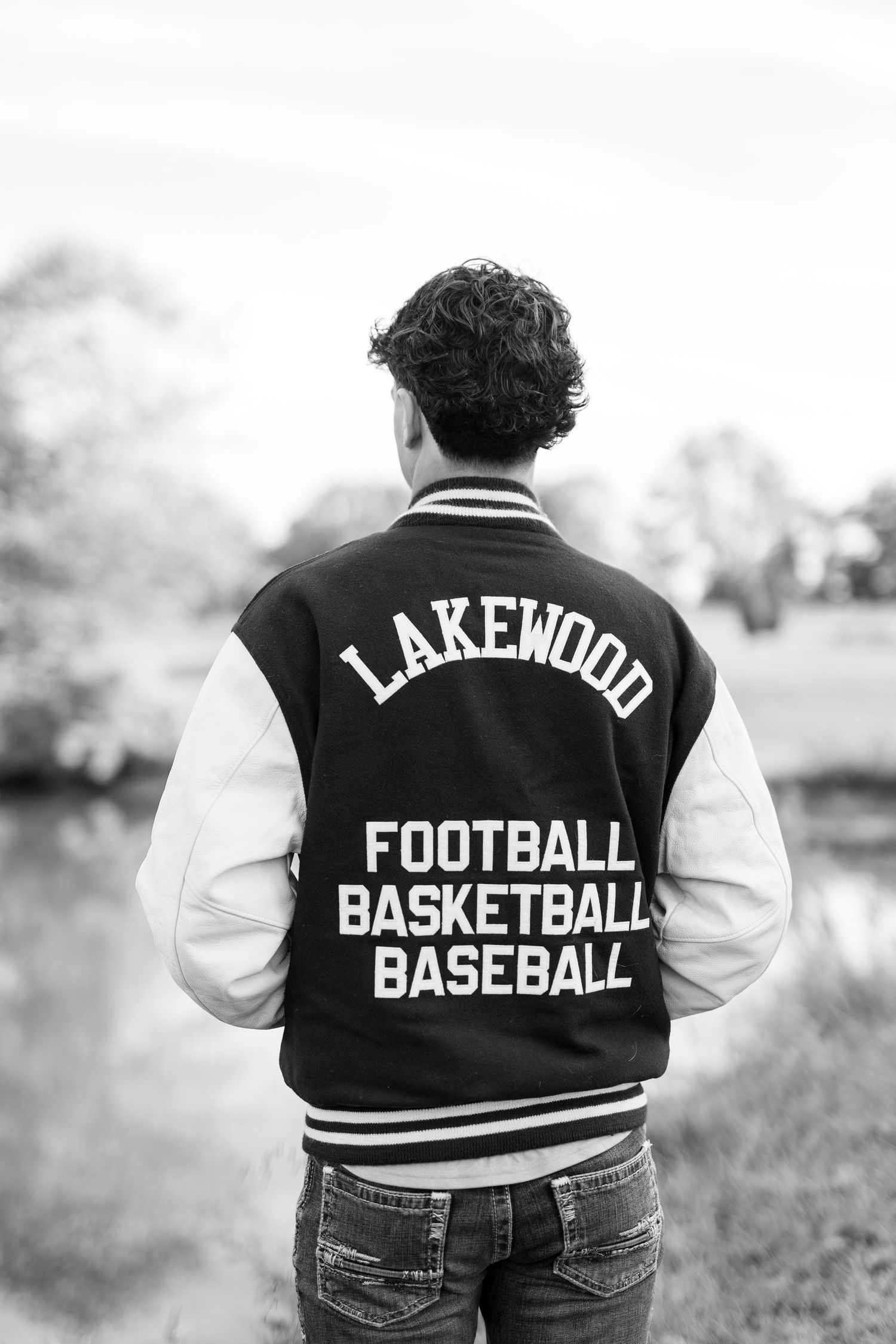 Black and white photo of a teen in a Lakewood varsity jacket listing football, basketball, and baseball, standing by a pond.