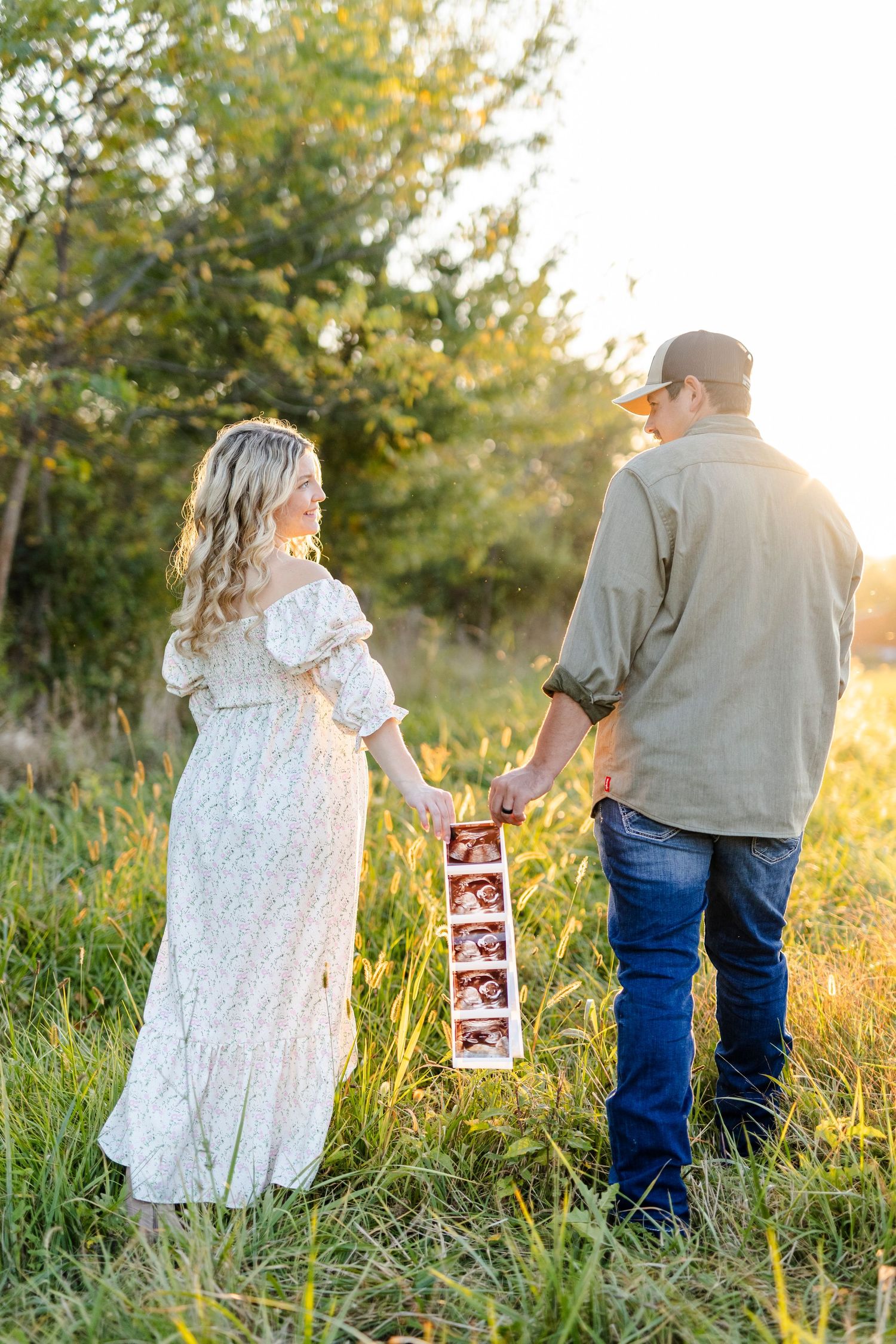 Couple holding hands and ultrasound photos in a sunlit field, maternity announcement portrait.