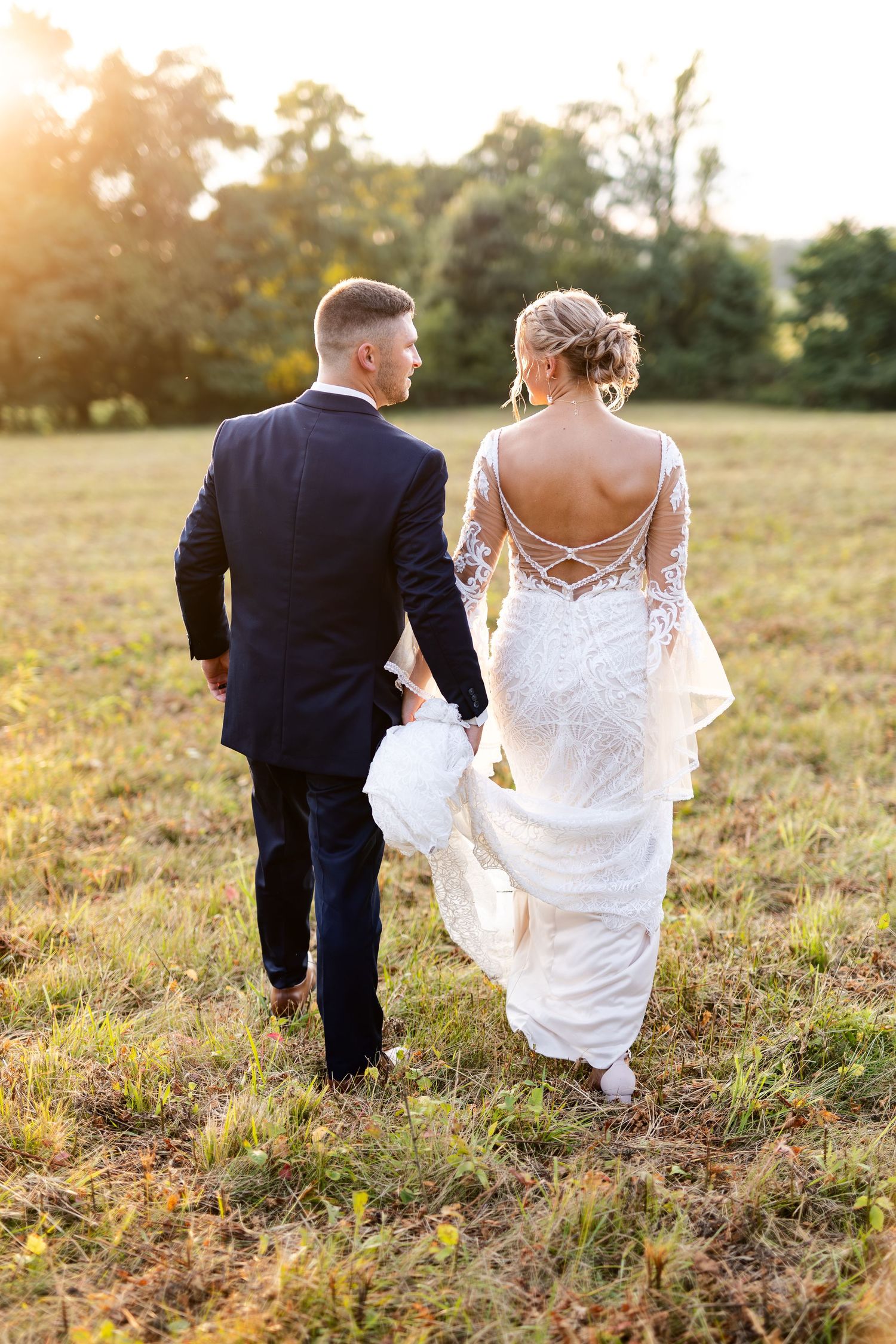 Bride and groom walk hand in hand away from camera through a golden sunset field.