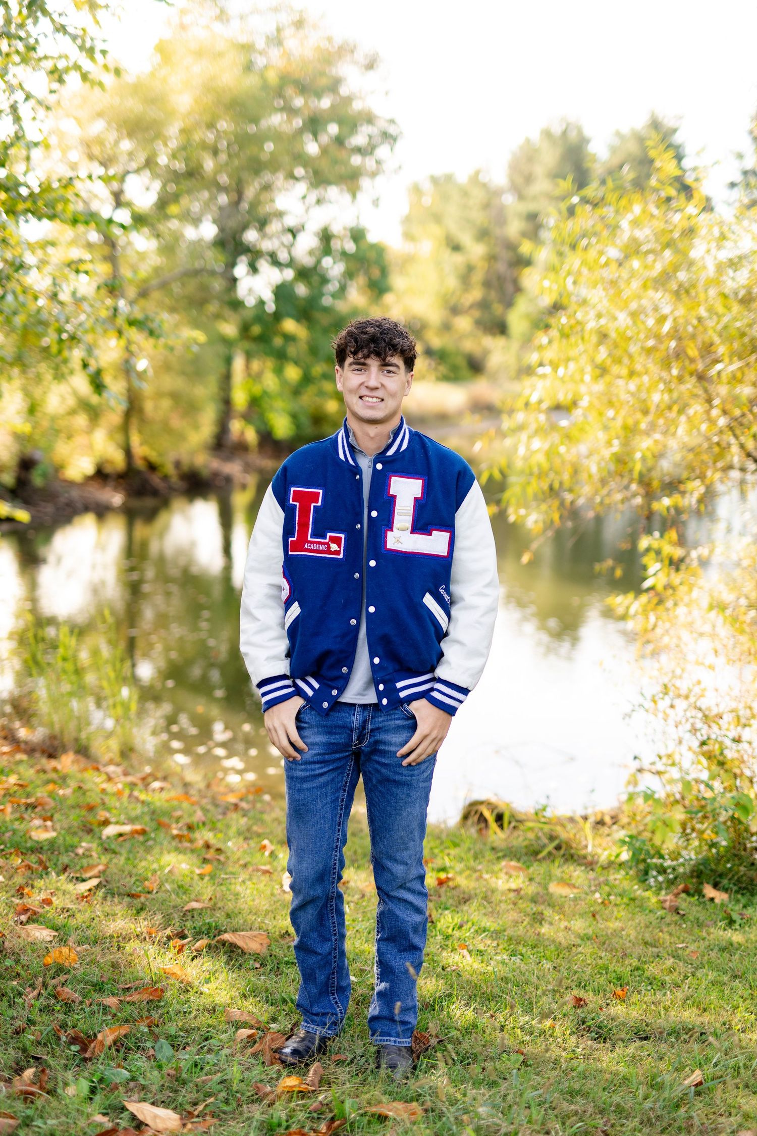 Young man in blue varsity letterman jacket with L letters smiling outdoors near pond in autumn park setting.